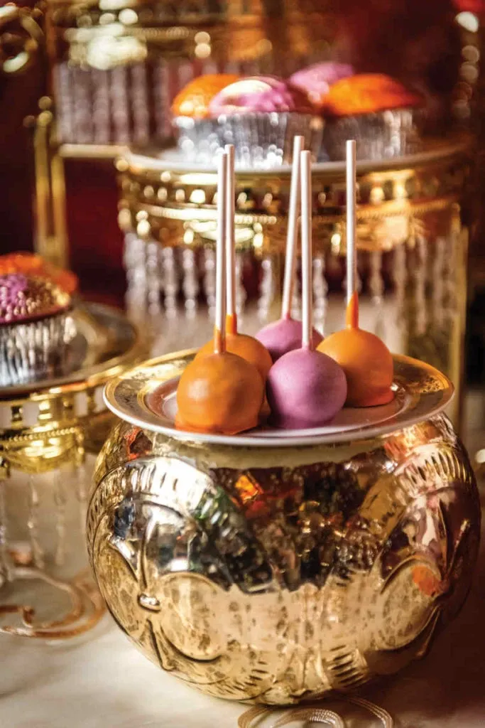 The image features a beautifully arranged display by Patisserie Dolci Piu, a vendor in the Wedding Cakes category. Five elegant cake pops, coated in vibrant shades of orange and pink, are artfully positioned on a decorative gold and white plate. The setup is elegantly situated on a gleaming, ornate gold stand. In the background, intricate decorations and shimmering glassware enhance the luxurious and festive atmosphere, ideal for an elegant wedding celebration.