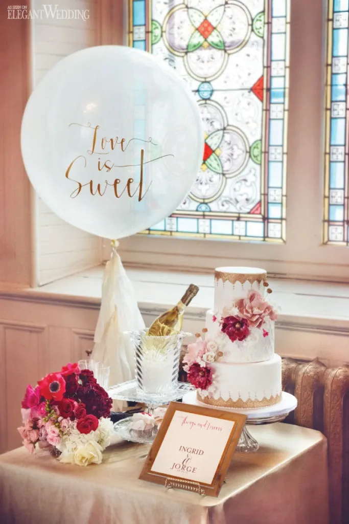 This image features a beautifully decorated wedding cake by Patisserie Dolci Piu, categorized under Wedding Cakes, displayed on a round table covered with an elegant white tablecloth. The cake is a two-tiered confection adorned with pink and white flower arrangements, showcasing intricate detailing. Beside the cake sits a vibrant bouquet of red and pink flowers, adding a pop of color to the setting. A bottle of champagne chilling in an ice bucket is also visible, suggesting celebratory toasts. A clear balloon with the phrase 'Love is Sweet' gently floats above, enhancing the romantic ambiance. In front of the cake, a small sign elegantly displays the names 'Ingrid and Jorge,' contributing to the personalized wedding theme. Richly colored stained glass windows in the background add a touch of timeless elegance to the scene.