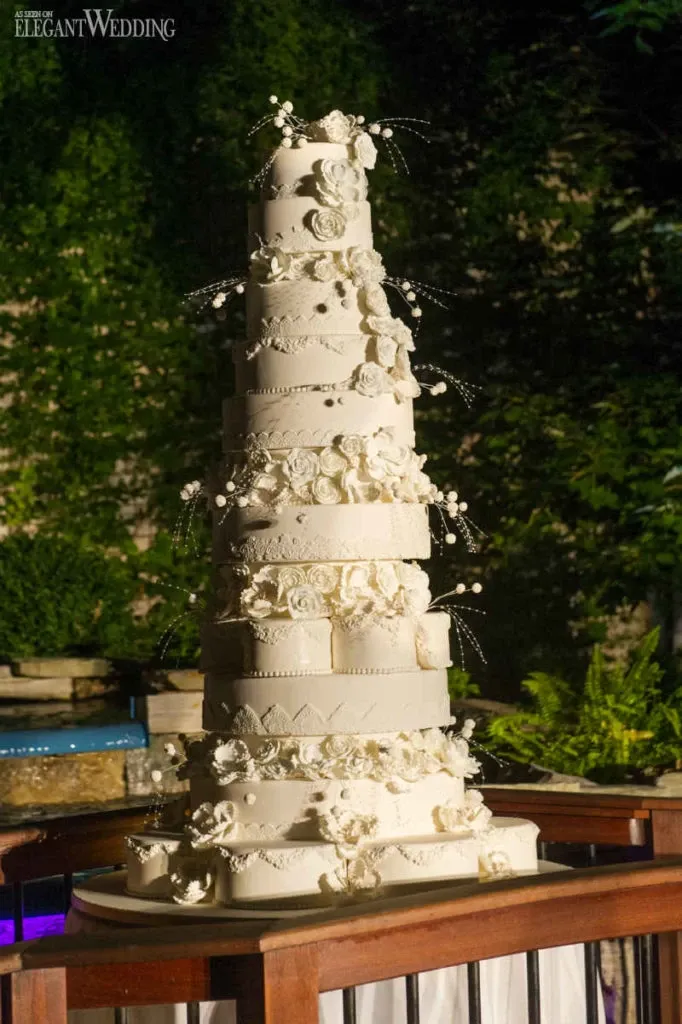 The image features an impressive, multi-tiered wedding cake crafted by Patisserie Dolci Piu, categorized under Wedding Cakes. This towering creation is elegantly adorned with intricate white fondant detailing and delicate sugar flowers cascading down the layers. Each tier exhibits ornate patterns, adding to the cake's sophistication. The backdrop includes lush greenery, suggesting an outdoor or garden wedding setting, complementing the cake's elegant appearance and enhancing its presentation for a wedding event hosted by Mariage Elegant in Canada.
