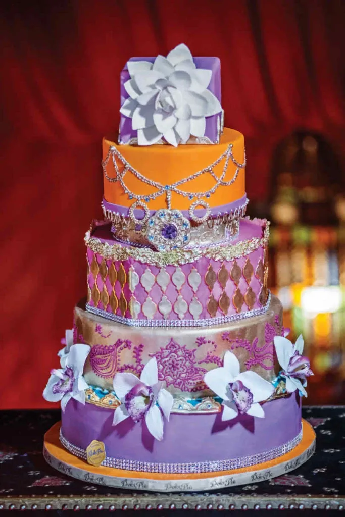 This image showcases an exquisite wedding cake created by Patisserie Dolci Piu, featured in the wedding cakes category on Mariage Elegant, Canada. The multi-tiered cake is a vibrant masterpiece with layers in shades of purple, orange, and gold. Each tier is adorned with intricate decorative elements, including a white floral arrangement sitting atop, delicate lace patterns on the middle tiers, and an elaborate jeweled design resembling a crown. The base is elegantly bordered with a shimmering ribbon, and the cake is embellished with white and purple flowers, contributing to its ornate and luxurious appearance. The cake stands against a rich red background, highlighting its grandeur and making it a perfect centerpiece for an elegant wedding celebration.
