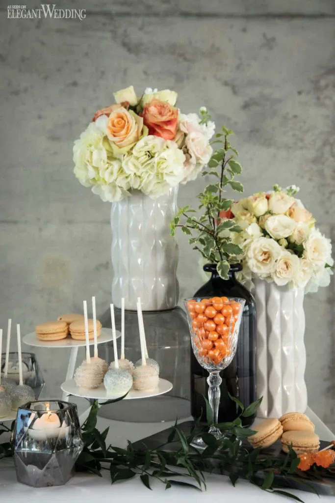 This elegant image showcases a display of exquisite wedding desserts by Patisserie Dolci Piu, categorized under Wedding Cakes for Mariage Elegant, Canada. The arrangement includes a selection of beautifully decorated cake pops and delicate macarons, artfully presented on tiered stands. The table is adorned with lush greenery and accented by a candle in a geometric holder, contributing to the sophisticated ambiance. Complementing the desserts are towering floral arrangements featuring cream and peach roses, which add a romantic touch to the setting. These elements come together to create a visually appealing and inviting display suitable for a wedding celebration.