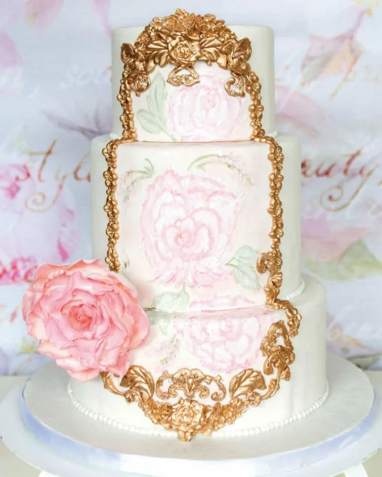 This image showcases an exquisite wedding cake made by Patisserie Dolci Piu, categorized under Wedding Cakes. The cake features three tiers, each adorned with intricate gold floral decorations and delicate pastel pink roses painted onto the surface. A large, elegant pink rose sits beautifully on the side of the bottom tier, adding a romantic touch. The cake is set against a soft, floral-patterned backdrop, enhancing its elegant and sophisticated appearance, perfect for a stylish wedding celebration.