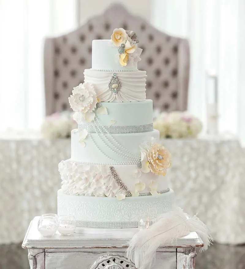 This image features an elegant wedding cake crafted by Patisserie Dolci Piu, categorized under Wedding Cakes, showcased on Mariage Elegant, Canada. The cake is a multi-tiered masterpiece adorned with intricate white icing patterns and cascading pearl-like details. Delicate white and soft yellow floral decorations add a touch of romance and sophistication. The middle tier is embellished with a silver brooch-like ornament. In the background, a plush tufted chair and a softly lit, elegantly decorated table set the scene for a luxurious wedding celebration.