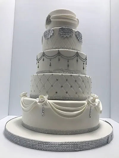 This image features an elegant, multi-tiered wedding cake created by Les Délices Lafrenaie, showcased under the wedding cakes category for Mariage Elegant in Canada. The cake is a luxurious five-tier design, adorned with intricate silver detailing that includes floral patterns and draped embellishments. The tiers display a quilted pattern with small beads, complementing the overall sophisticated aesthetic. Each layer is separated by sparkling silver bands, and the top tier is decorated with a hat-like topper. This cake epitomizes a refined and glamorous style, perfect for a grand wedding celebration.