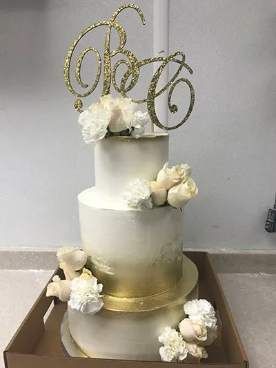This image features a stunning three-tiered wedding cake by Les Délices Lafrenaie, categorized under Wedding Cakes for Mariage Elegant, Canada. The cake is elegantly frosted in a smooth, white icing with subtle gold accents encircling the base of each tier. Adorning the cake are delicate white roses and peonies, adding a touch of floral charm. The top tier is crowned with a sophisticated gold monogram cake topper, perfectly complementing the luxe theme. The cake, displayed in a classic setting, exemplifies elegance and is ideal for a formal wedding celebration.