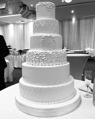 This image showcases an elegant, multi-tiered wedding cake crafted by Les Délices Lafrenaie, featured in the Wedding Cakes category. The cake is beautifully decorated with intricate patterns and floral embellishments, demonstrating exceptional artistry and attention to detail. Set in a sophisticated reception venue with draped curtains and ambient lighting, the scene highlights the cake's central role in a wedding celebration. A champagne flute is visible on the side, symbolizing celebratory toasts and the joyous atmosphere of a wedding event.