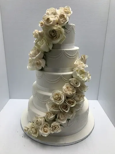 This image shows a stunning five-tier wedding cake created by Les Délices Lafrenaie, categorized under Wedding Cakes for Mariage Elegant, Canada. The cake is elegantly adorned with cascading white roses, intricately placed from the top tier to the base. Each tier is beautifully decorated with delicate pearl-like icing patterns, adding a touch of sophistication. The classic white design and floral arrangement make it a perfect centerpiece for an elegant wedding celebration.