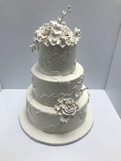 This image features a stunning three-tiered wedding cake by Les Délices Lafrenaie, showcased under the Wedding Cakes category on Mariage Elegant. The elegantly designed cake is adorned with intricate white floral motifs and delicately crafted sugar flowers that beautifully cascade from the top tier. The cake's subtle elegance reflects classic wedding sophistication, making it an ideal choice for those seeking a timeless centerpiece for their special day. The background is simple, ensuring full focus on the cake's exquisite detailing.