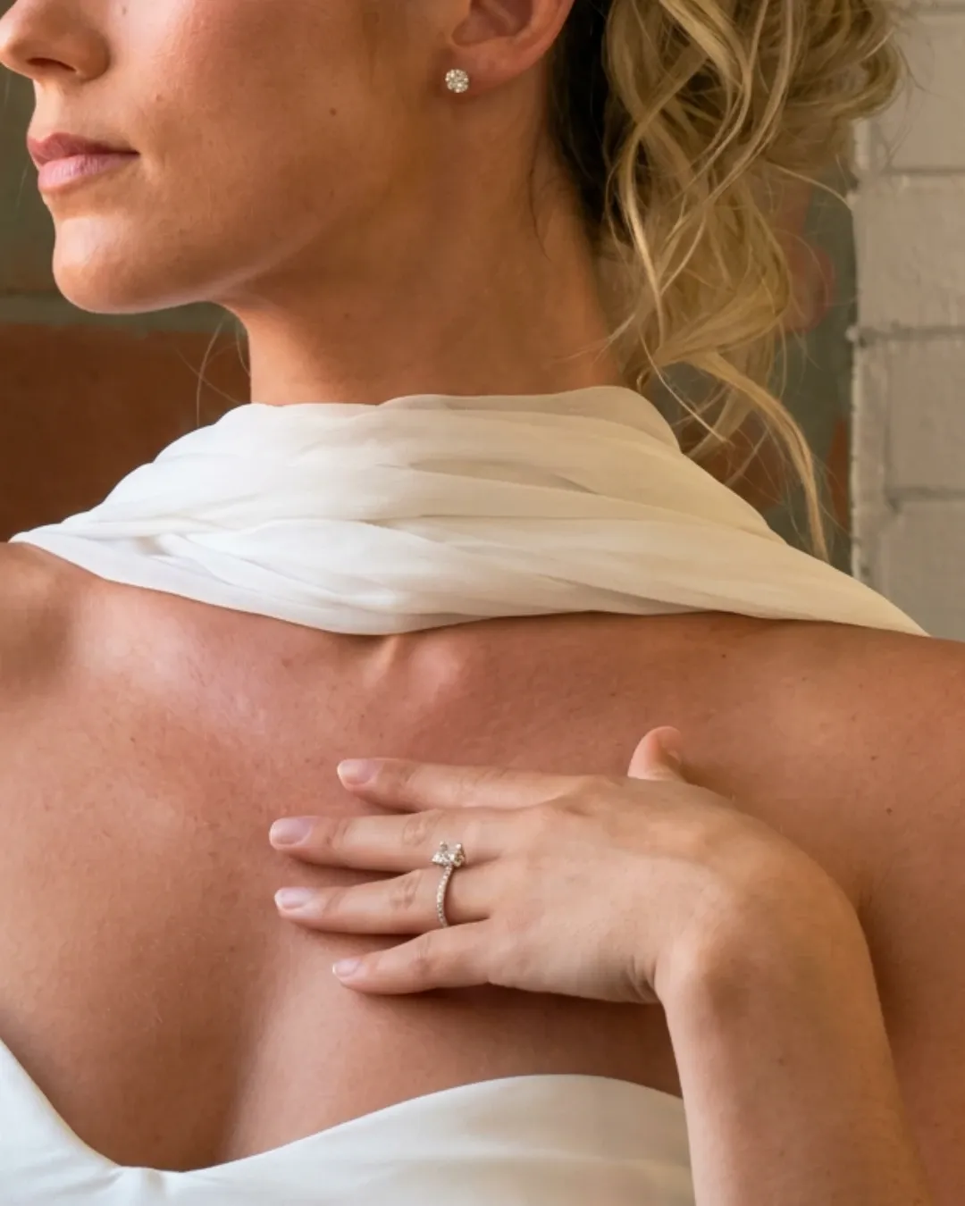 A woman is elegantly posed with her back to the camera, highlighting a sophisticated updo hairstyle. She is adorned with a delicate pair of stud earrings and wears a luxurious white scarf draped gracefully around her shoulders. Her fingers rest gently on her shoulder, showcasing a stunning diamond engagement ring. The setting suggests a focus on bridal jewelry, perfect for wedding ceremonies. The image captures the essence of elegance and sophistication, making it an ideal representation for a wedding jewelry section on the Mariage Elegant website in Canada.