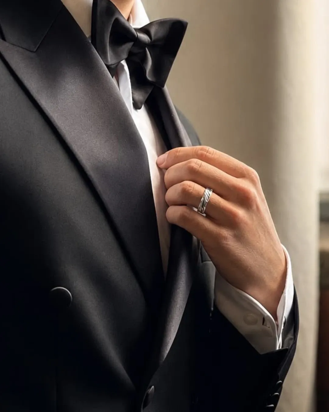 A person dressed in a formal black tuxedo is adjusting the lapel with their left hand, showcasing a polished wedding band on their ring finger. The tuxedo features a satin bow tie and a crisp white dress shirt, suggesting an elegant setting suitable for a wedding. The focus on the ring emphasizes its significance as wedding jewelry, making it an ideal representation for Mariage Elegant's wedding vendor listings in Canada.