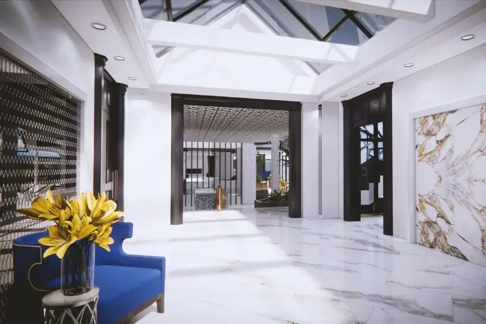 The image showcases a luxurious and modern wedding venue interior, featuring a spacious area with gleaming white marble flooring and elegant architectural details. The ceiling includes expansive glass skylights, allowing natural light to flood the space. On the left side, a vibrant blue chair and a vase filled with yellow flowers add a pop of color and sophistication. A striking wall with intricate patterns complements the overall classy ambiance. This venue offers a stylish and inviting setting for weddings, located in Canada and is listed on Mariage Elegant.