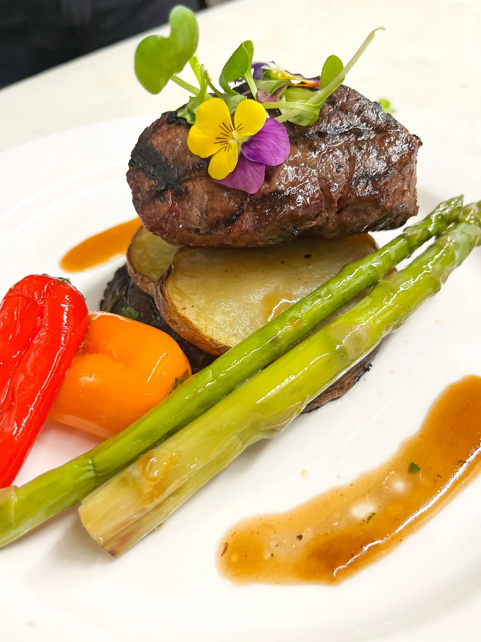 A beautifully plated wedding dish featuring a succulent grilled steak atop a stack of roasted potato slices. The dish is artfully garnished with vibrant edible flowers and fresh microgreens, adding a touch of elegance. Accompanying the steak are perfectly cooked asparagus spears and colorful bell peppers, drizzled with a rich, savory sauce. Ideal for upscale wedding receptions, this gourmet presentation showcases the culinary expertise available at premium wedding venues listed on Mariage Elegant, Canada.