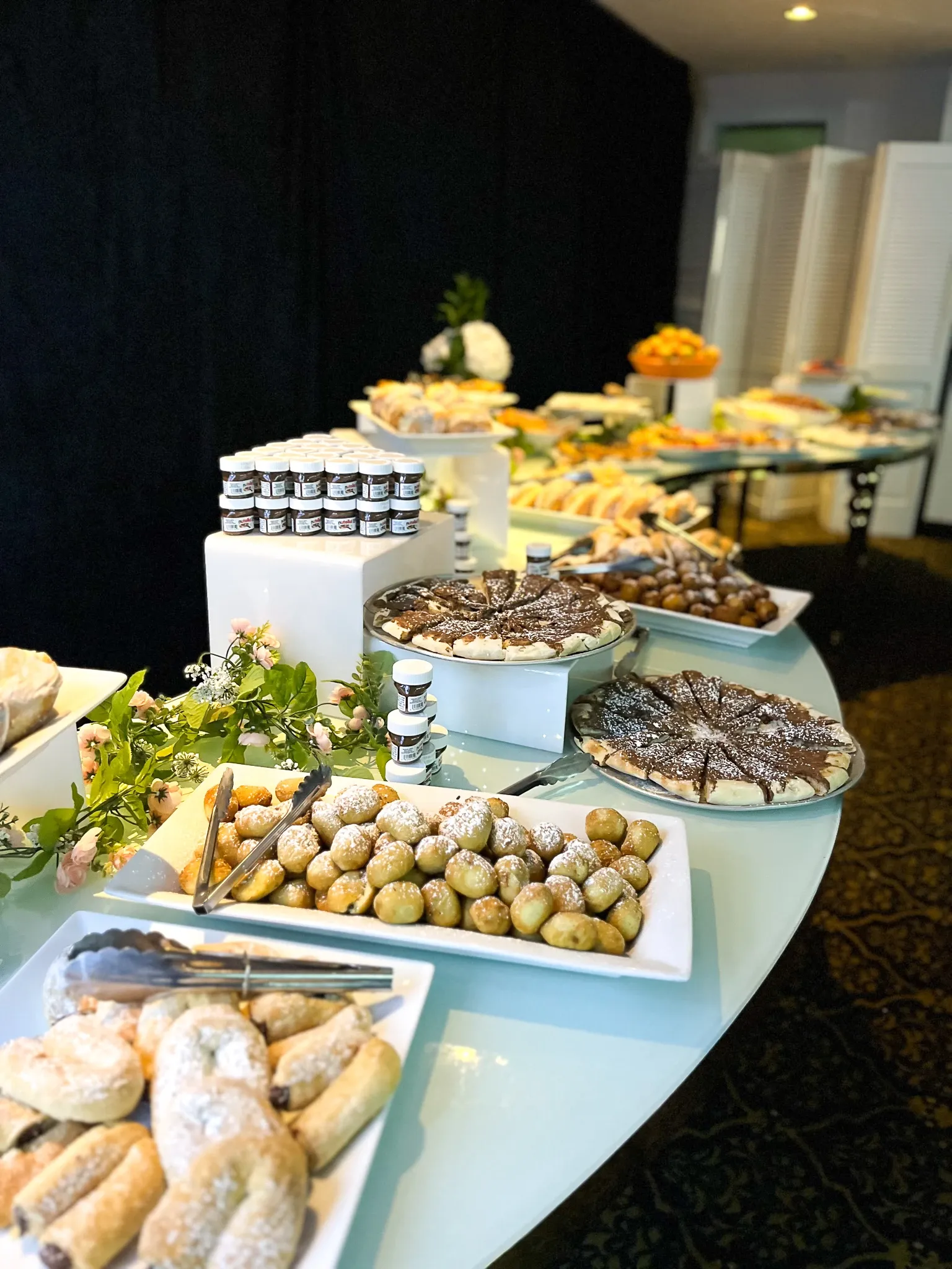 The image features an elegantly arranged dessert table at a wedding venue, showcasing a variety of sweets and pastries. In the foreground, there are plates of pastries dusted with powdered sugar, alongside a selection of chocolate-topped desserts. A decorative sprig of greenery adds a touch of freshness to the display. In the background, several plates with assorted bite-sized treats and two jars of honey or jam are visible. The table, covered in a sleek white tablecloth, suggests a sophisticated and inviting atmosphere typical of upscale wedding venues.