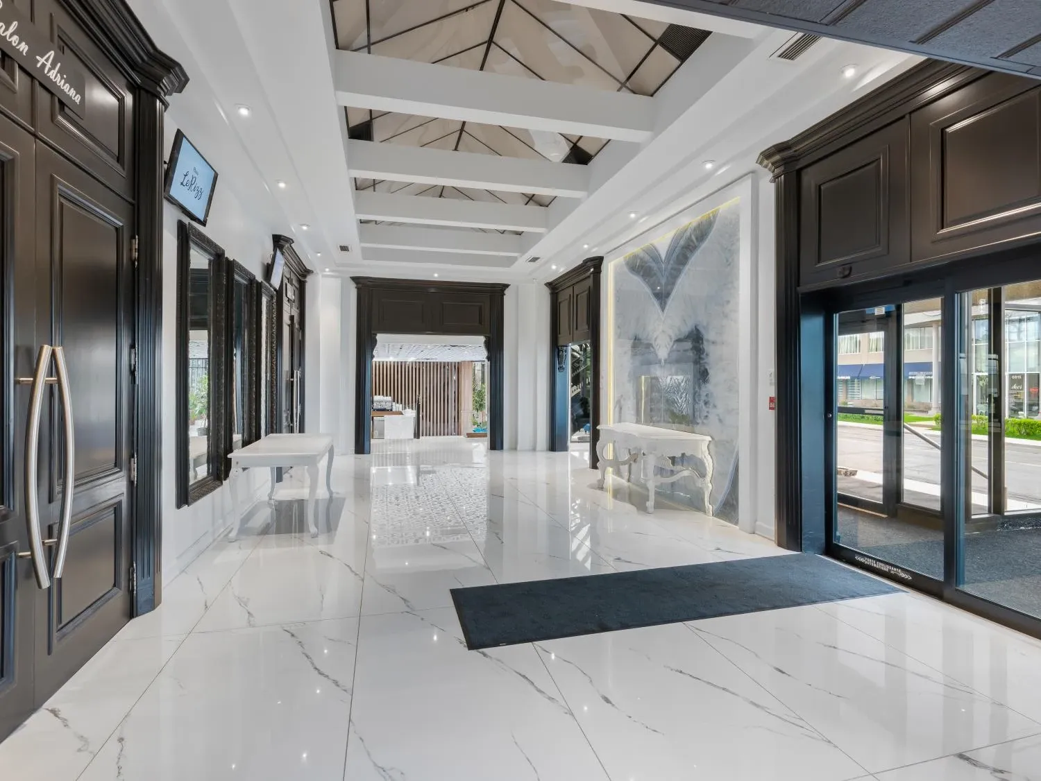 La photo montre une élégante salle de réception avec un sol en marbre blanc brillant. Des murs aux moulures noires encadrent l'espace, tandis qu'un plafond haut et voûté, décoré de détails sophistiqués, ajoute de la grandeur à l'ensemble. Une grande porte vitrée sur le côté droit laisse entrer la lumière naturelle. À gauche, un panneau signalétique stylisé est visible, probablement pour indiquer le lieu ou l'événement. Tout au fond, une table décorative blanche est placée contre un mur orné d'une large œuvre d'art abstraite. Cette salle de réception, idéale pour un mariage, combine luxe contemporain et charme classique, offrant un cadre raffiné pour les célébrations nuptiales.