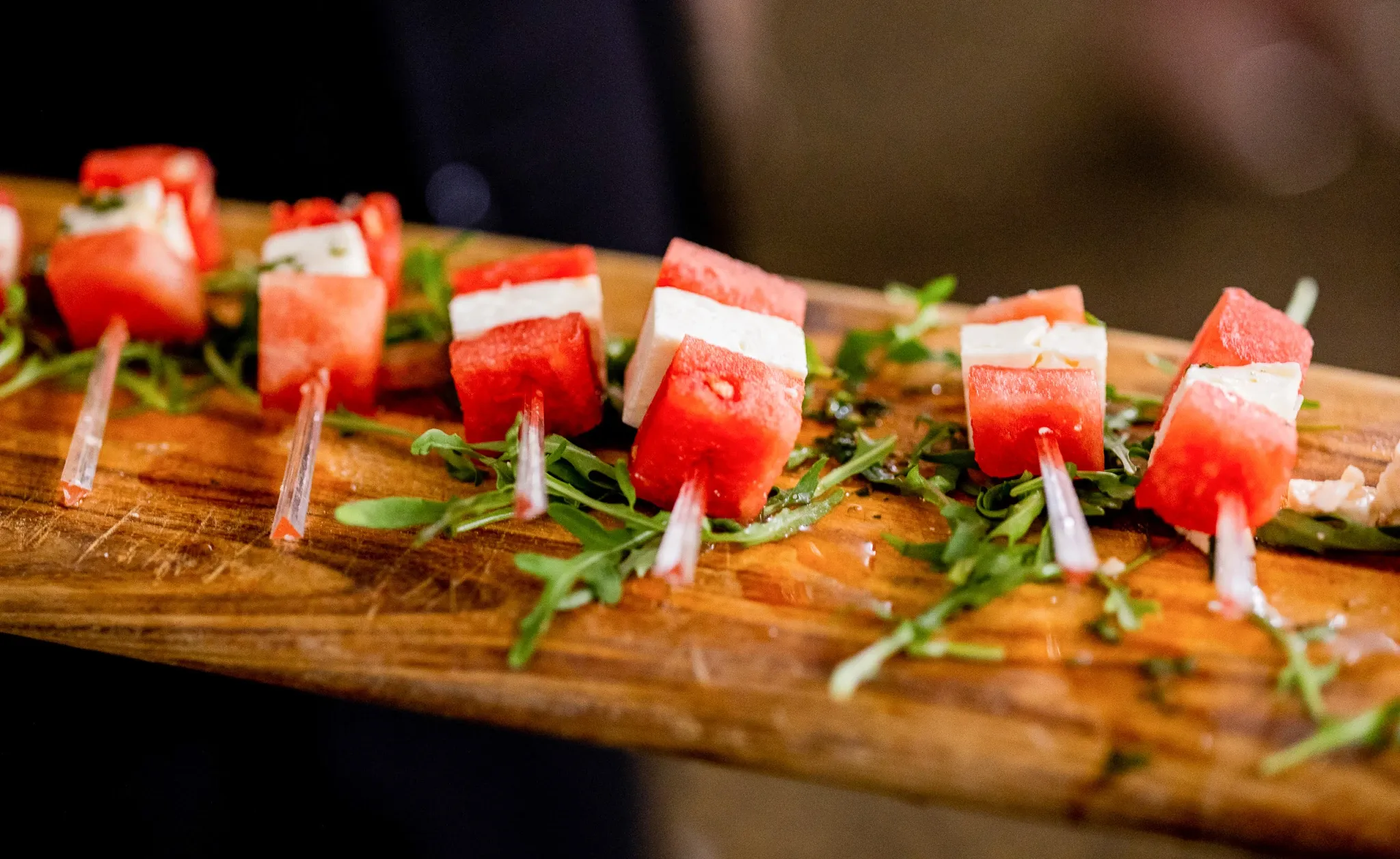 Une planche en bois élégante présente des amuse-bouches sophistiqués, composés de cubes de pastèque juteuse et de fromage feta, délicatement enfilés sur des piques en plastique transparent. Les brochettes sont disposées sur un lit de roquette fraîche, ajoutant une touche de couleur verte vibrante au plat. Cet assortiment gastronomique est idéal pour un cocktail de mariage raffiné, mettant en valeur l'attention au détail et la qualité des services traiteurs proposés par Mariage Elegant, Canada.