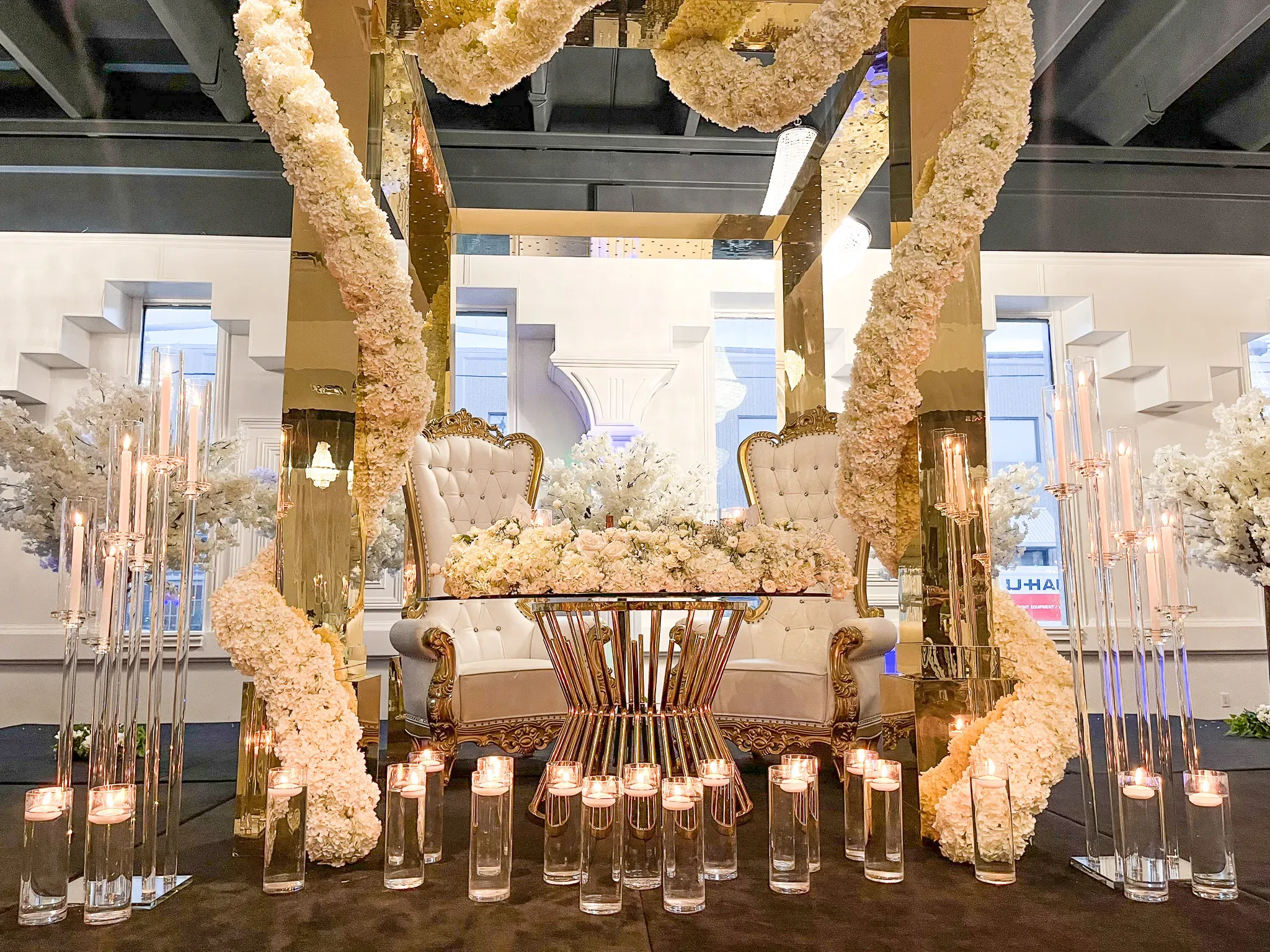A lavish wedding venue showcasing an ornately decorated setting. At the center is an elegant throne-style loveseat draped in luxurious white fabric, surrounded by an exquisite floral arrangement featuring cascading white flowers. The backdrop includes tall, gold-accented mirrors that enhance the grandeur of the setup. Flanking the arrangement are sleek glass candleholders with tall, slender candles, adding a romantic ambiance. The overall sophistication of the decor reflects an upscale venue perfect for a luxurious wedding celebration.