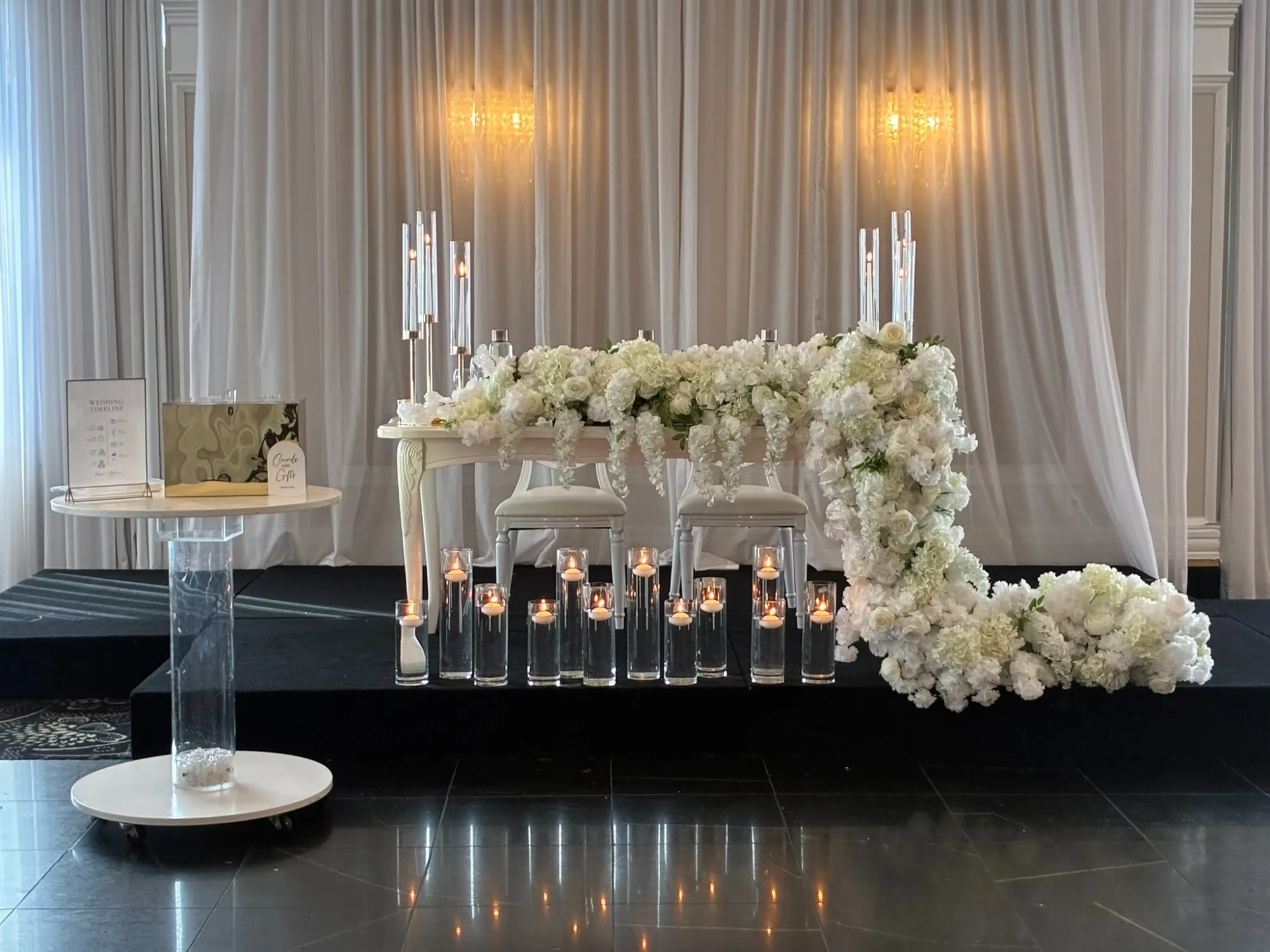 The image showcases a beautifully decorated wedding venue by Plaza Universel, categorized under Wedding Venues at Mariage Elegant, Canada. The setting includes an elegant table adorned with a lush arrangement of white flowers cascading over the edge. Behind the table, sheer white drapes create a soft, romantic backdrop, enhanced by the warm glow of overhead chandeliers. In front of the table, numerous tall glass vases with flickering candles add a touch of sophistication and ambiance to the scene. To the side, a small pedestal table holds a guest book, emphasizing the venue's attention to detail and readiness for a memorable wedding celebration.