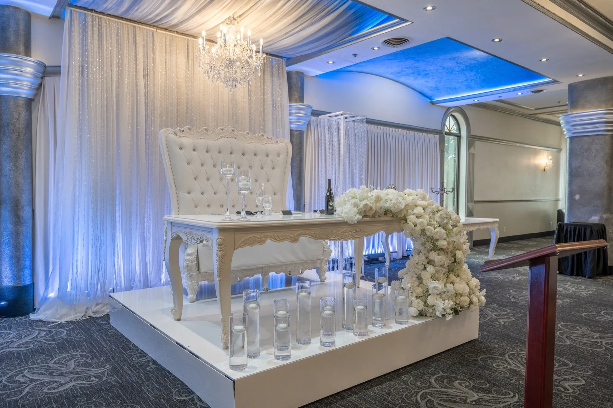 The image showcases a beautifully decorated wedding venue at Plaza Marina, part of the wedding venues category on Mariage Elegant, Canada. The setting features an elegant white table adorned with a lush floral arrangement cascading over the edge. Behind the table stands a luxurious high-backed tufted chair. The background is a shimmering, light-colored drapery with a sparkling chandelier hanging above, adding an air of sophistication. The ceiling is accented with soft blue lighting, enhancing the romantic ambiance of the venue. Candles arranged in clear glass holders surround the table, contributing to the serene and opulent atmosphere.