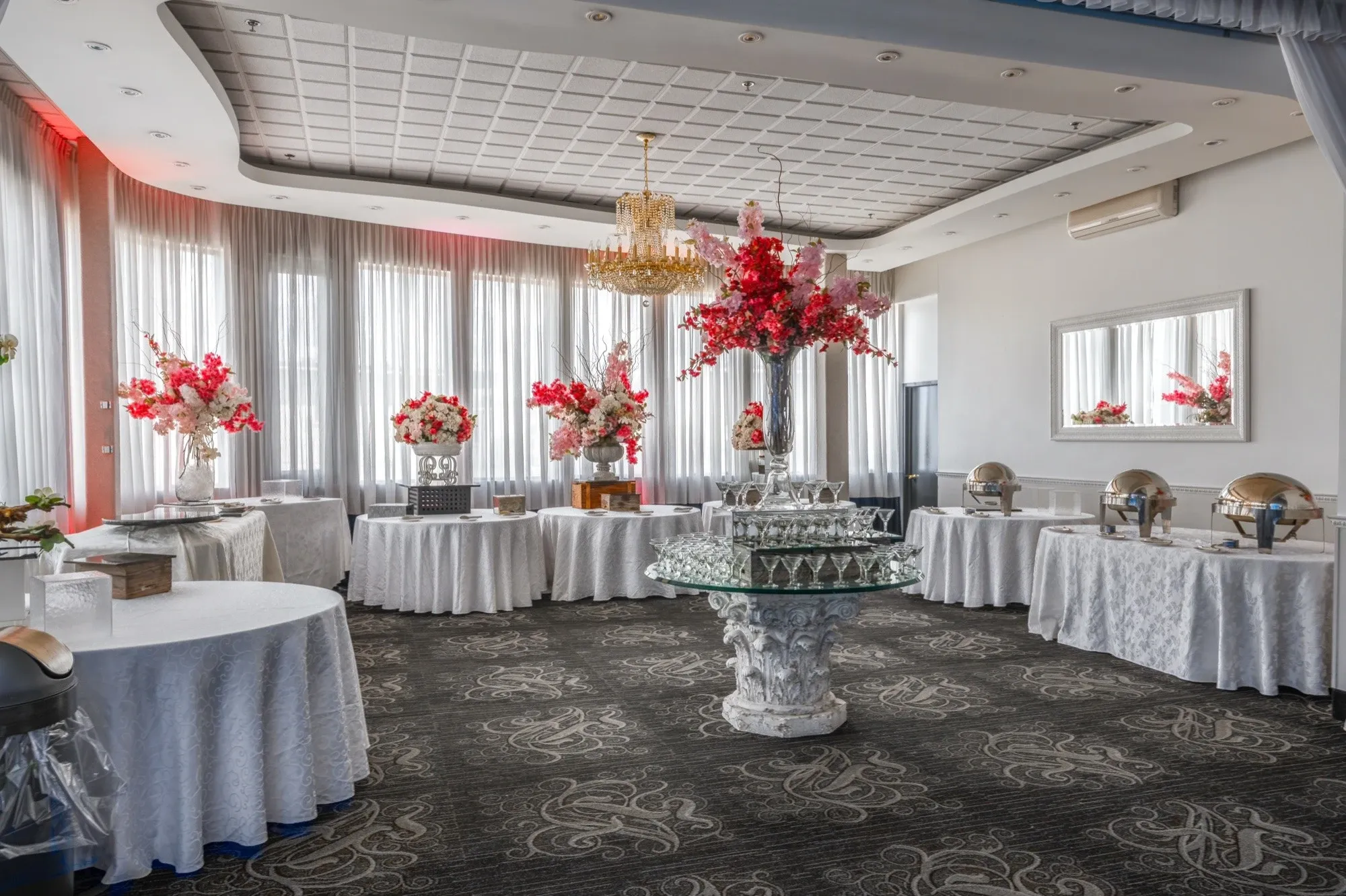 The image showcases a beautifully decorated wedding venue at Plaza Marina, highlighting its elegance for Mariage Elegant's wedding venue category in Canada. The spacious room features a series of round tables covered with white tablecloths, each adorned with striking floral arrangements in vibrant red and white hues. A lavish chandelier hangs from the ceiling, adding a touch of opulence. Large windows with sheer curtains allow ample natural light, creating a bright and inviting atmosphere. The setup includes an ornate central table with a glass display, emphasizing the sophisticated ambiance suitable for weddings and receptions.