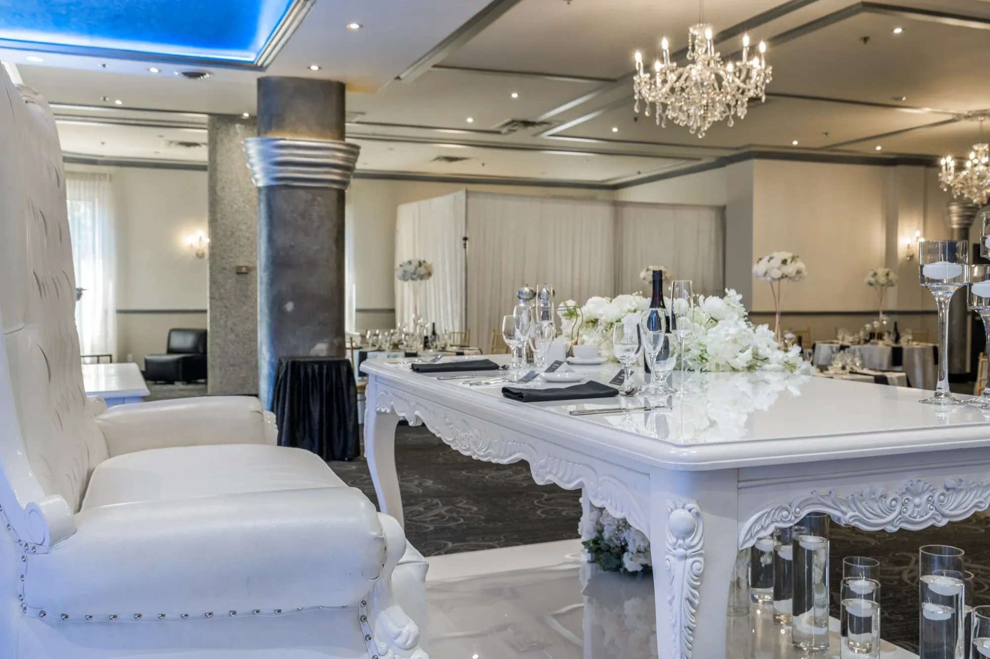 The image features an elegantly decorated wedding venue interior at Plaza Marina, a prominent wedding venue listed on Mariage Elegant in Canada. A lavish, white table is beautifully set with crystal wine glasses, intricately folded napkins, and an array of elegant silverware. A luxurious, white upholstered chair is prominently placed at one end of the table. The table is adorned with a lush arrangement of white flowers, complementing the sophisticated setting. Overhead, sparkling chandeliers illuminate the room, adding a touch of grandeur. The venue's chic decor, including the sleek gray pillar and ambient lighting, creates an inviting atmosphere ideal for wedding celebrations.