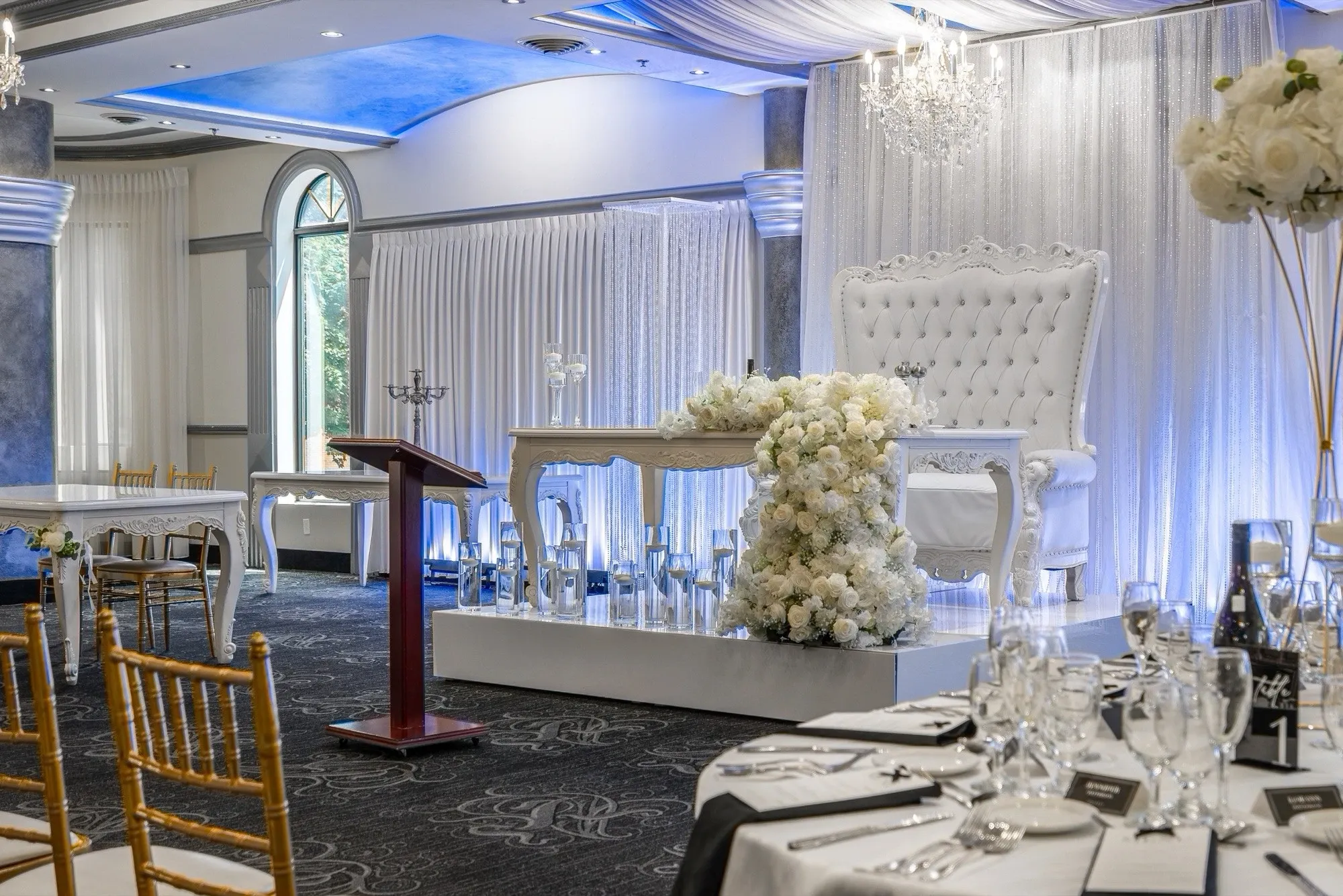 The image showcases an elegant wedding venue setup at Plaza Marina, categorized under Wedding Venues for Mariage Elegant in Canada. Prominently featured is a luxurious reception area adorned with a majestic white throne-style chair on a raised platform, decorated with cascading white flowers. The background showcases shimmering silver drapes complementing the opulent chandelier above. A wooden podium is positioned nearby, suggesting a space for speeches or announcements. The tables are elegantly set with fine glassware, neatly folded napkins, and floral centerpieces, offering a sophisticated ambience suitable for weddings. The venue's high ceilings and large arched windows provide natural light, enhancing the overall elegance of the space.