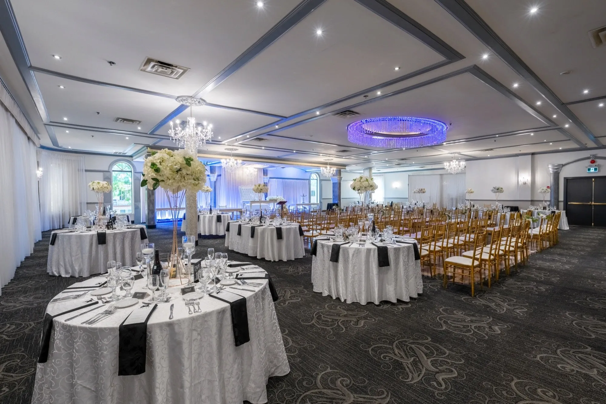 The image showcases the interior of Plaza Marina, an elegant wedding venue under the wedding venues category, featured on Mariage Elegant, Canada. The spacious room is beautifully arranged for a wedding reception with round tables draped in white linens and adorned with tall floral centerpieces comprising white blooms. Gold chiavari chairs are neatly aligned both around the tables and in rows, suggesting a versatile setup accommodating both dining and ceremony seating. The ceiling features luxurious chandeliers and a striking purple LED-lit circular piece, enhancing the venue's sophisticated ambiance. Soft lighting and large arched windows in the background add to the venue’s inviting atmosphere, ideal for celebrating special occasions.