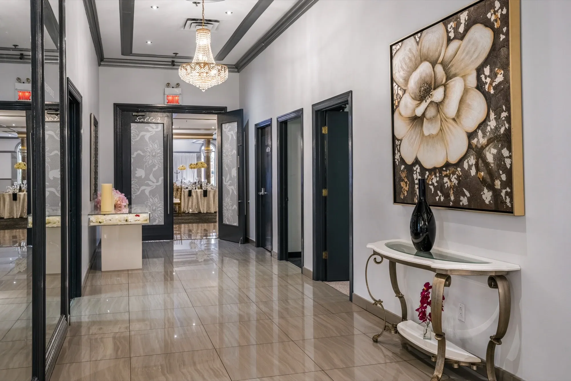 The image features an elegant hallway at Plaza Marina, a premier wedding venue listed on Mariage Elegant, Canada. The hallway is adorned with a shimmering chandelier that casts a gentle glow on the polished tile floor. A striking floral artwork decorates the wall, providing an artistic touch. On the right, a decorative table holds a sleek black vase, adding a modern element to the sophisticated ambiance. Beyond the hallway, open doors reveal a glimpse of a beautifully set wedding reception area, indicating the venue's readiness for a stylish celebration.