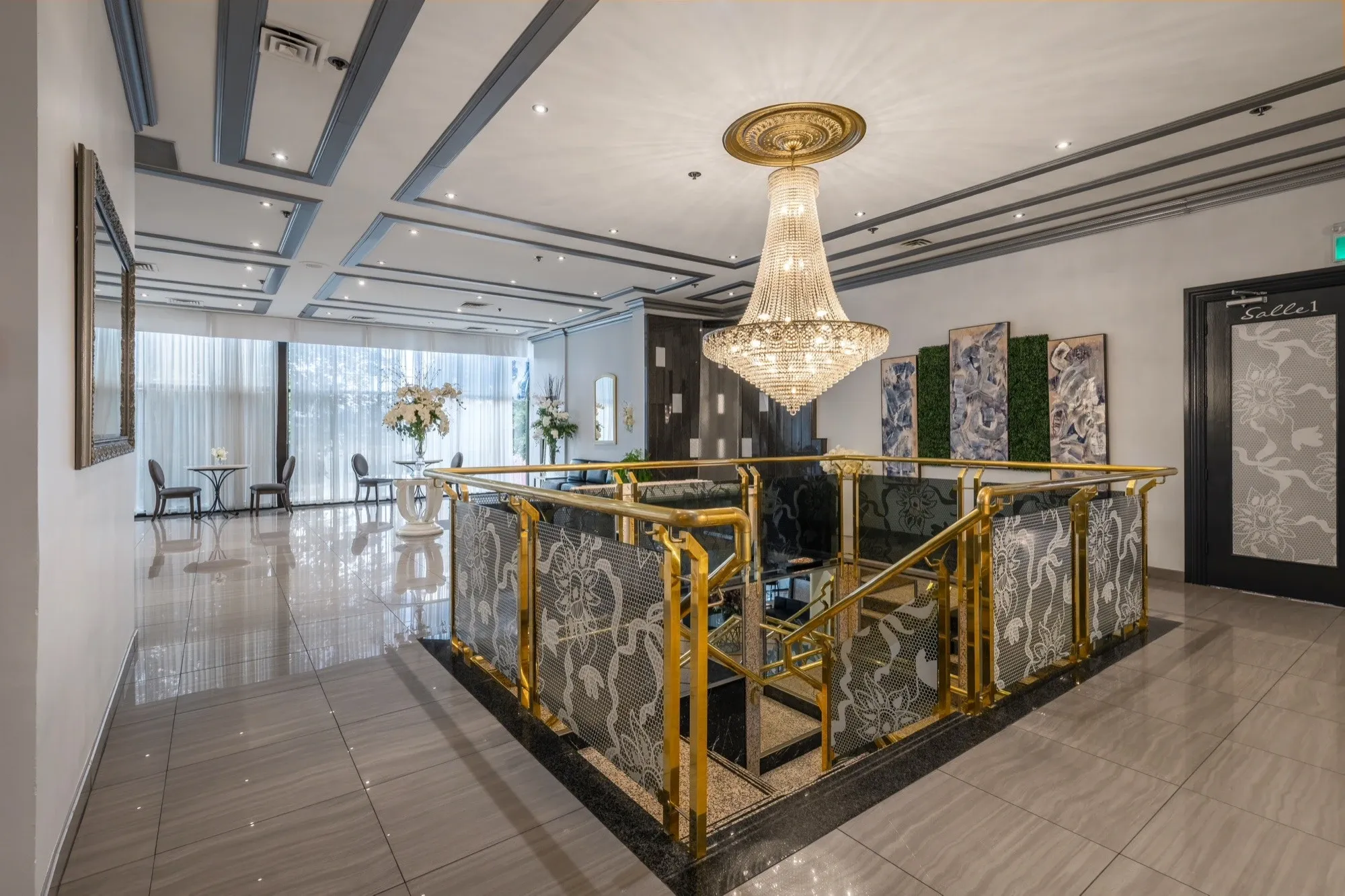 The image showcases Plaza Marina, a premier wedding venue featured on Mariage Elegant, Canada. The venue's interior is elegantly designed, featuring a luxurious chandelier hanging above a staircase with ornate, golden railings. The surrounding walls are adorned with intricate art pieces, creating a sophisticated atmosphere. Large windows to the left allow natural light to flood the space, highlighting tables arranged with stylish floral centerpieces, enhancing the venue's romantic appeal for weddings and special events.