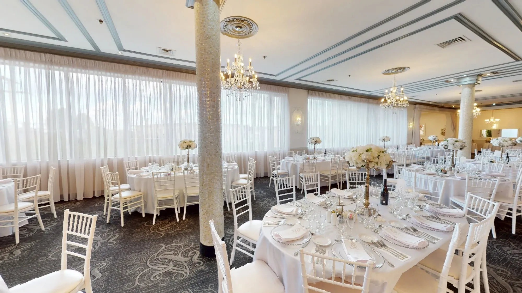 The image showcases the Plaza Marina, a featured wedding venue on Mariage Elegant, Canada. The venue's interior is elegantly decorated for a wedding reception, with round tables set for guests, adorned with white tablecloths and floral centerpieces. The room is illuminated by crystal chandeliers that hang from the ceiling, adding a touch of sophistication. White Chiavari chairs are neatly arranged around the tables, and the large windows draped with sheer curtains allow natural light to fill the space. The venue's refined atmosphere is ideal for hosting elegant wedding celebrations.