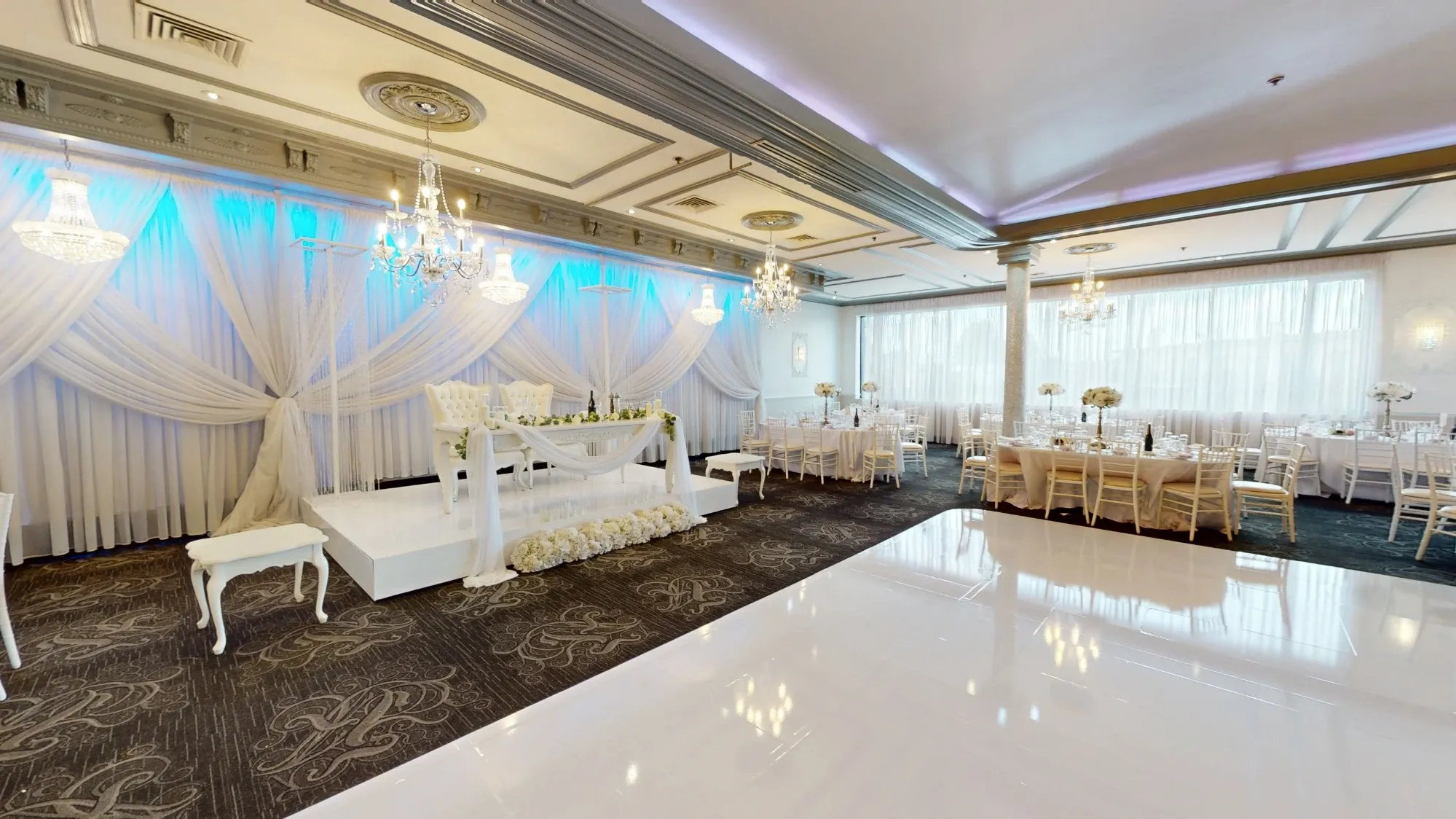 This image showcases Plaza Marina, featured in the Wedding Venues category on Mariage Elegant, Canada. The venue is elegantly decorated with luxurious chandeliers hanging from an ornate ceiling. The focal point is a beautifully draped stage adorned with white flowers, perfect for wedding ceremonies. Surrounding the stage are tables set for a formal reception, covered with white tablecloths and embellished with floral centerpieces. Soft, ambient lighting highlights the sophisticated decor, creating an inviting and romantic atmosphere ideal for weddings. A polished white dance floor is in the foreground, ready for guests to celebrate.