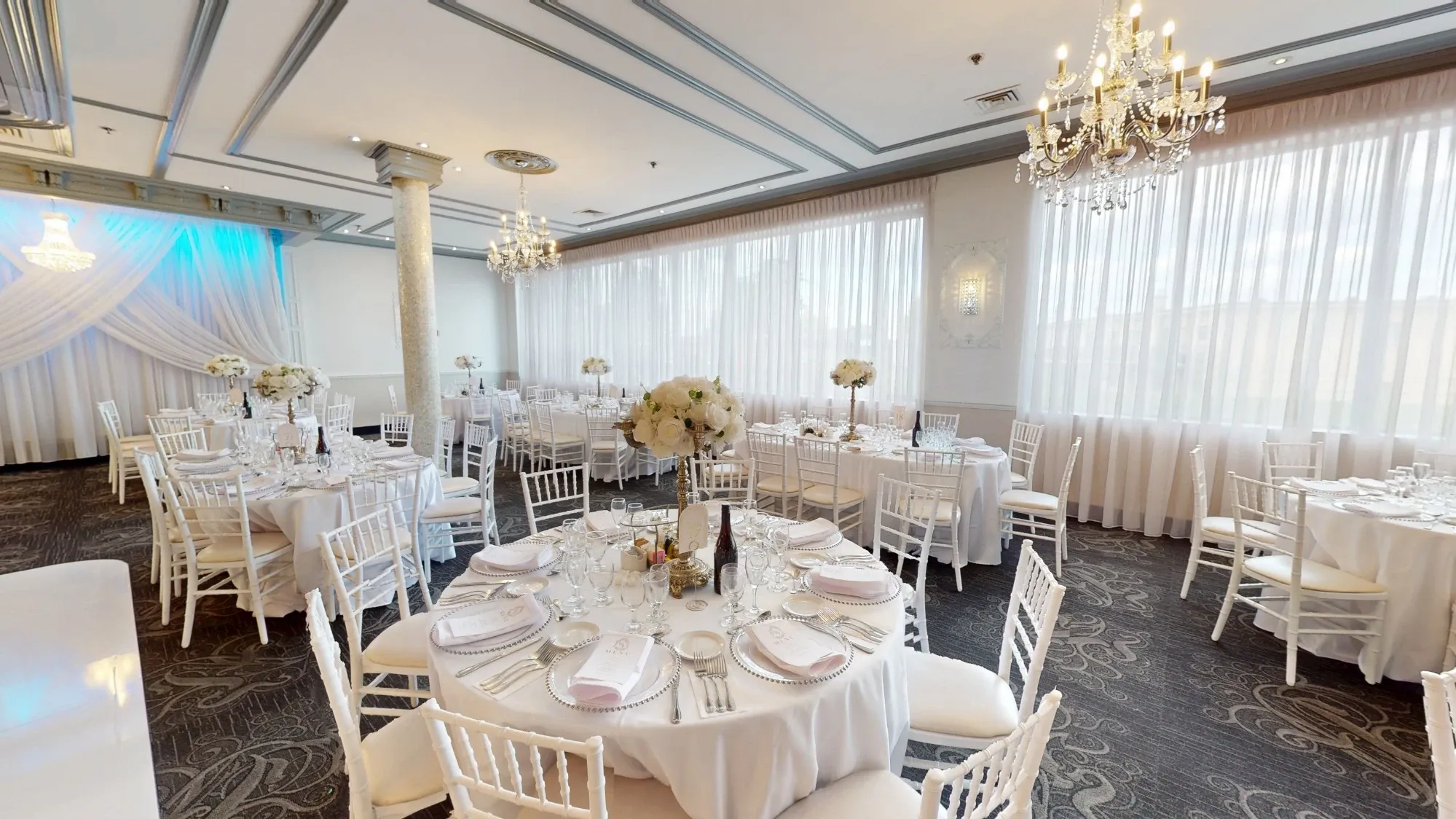 The image shows the elegant interior of Plaza Marina, a premier wedding venue listed under Mariage Elegant, Canada. The room features several round tables covered with pristine white tablecloths, each adorned with centerpieces of white flowers. Crystal chandeliers hang from the ceiling, providing a soft, ambient glow that complements the natural light streaming in through large, sheer-curtained windows. White chairs are neatly arranged around the tables, all set with fine china and glassware, creating a sophisticated atmosphere perfect for a wedding reception.