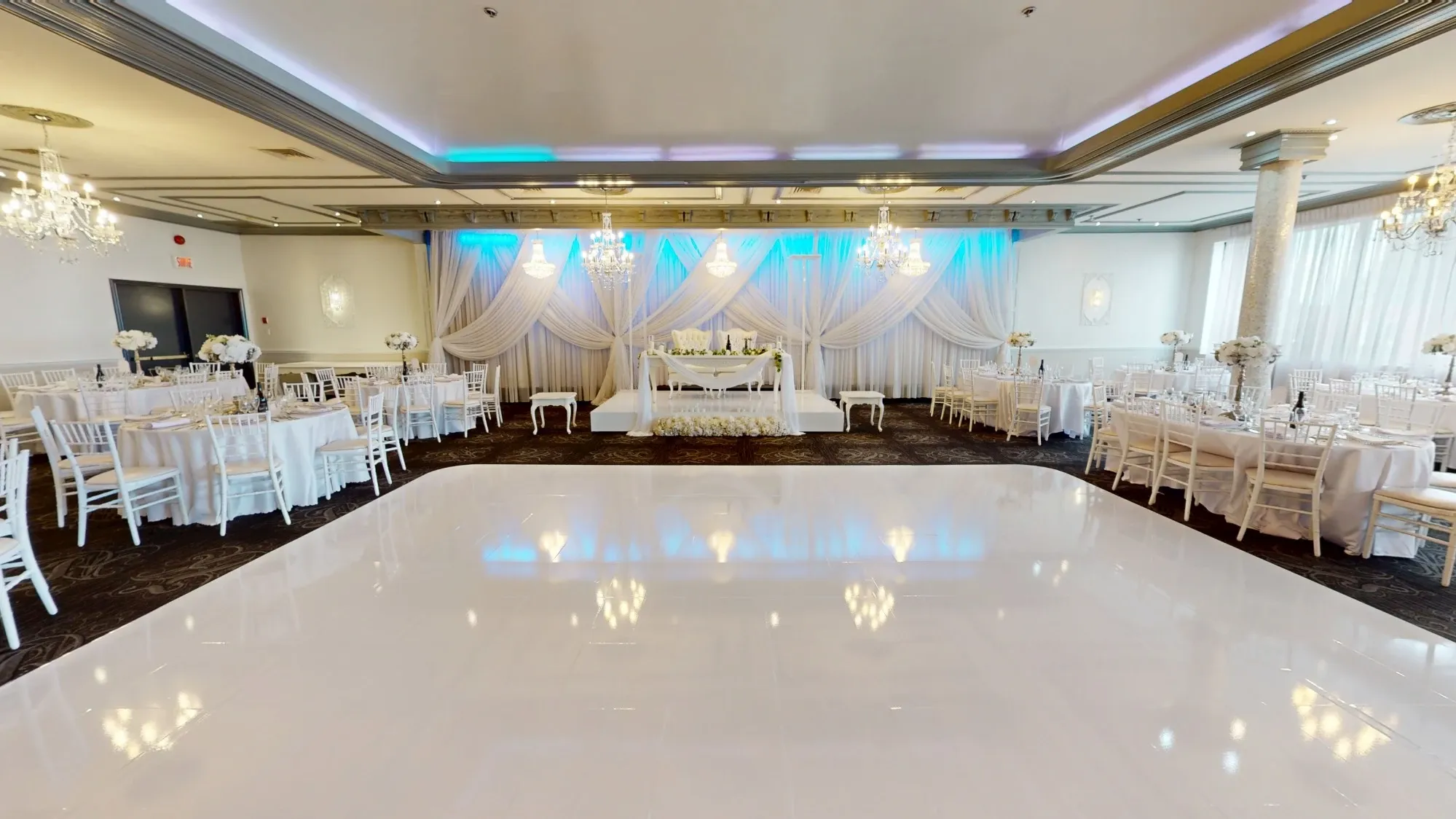 The image showcases an elegant wedding venue at Plaza Marina, a featured vendor on Mariage Elegant, Canada, under the Wedding Venues category. The spacious hall is adorned with luxurious chandeliers casting a warm glow over the room. White drapes elegantly frame a tastefully set head table at the center, complemented by blue ambient lighting. Round tables are neatly arranged around the polished dance floor, each set with white chairs and adorned with simple, yet stylish floral centerpieces, setting a sophisticated ambiance perfect for a wedding celebration.