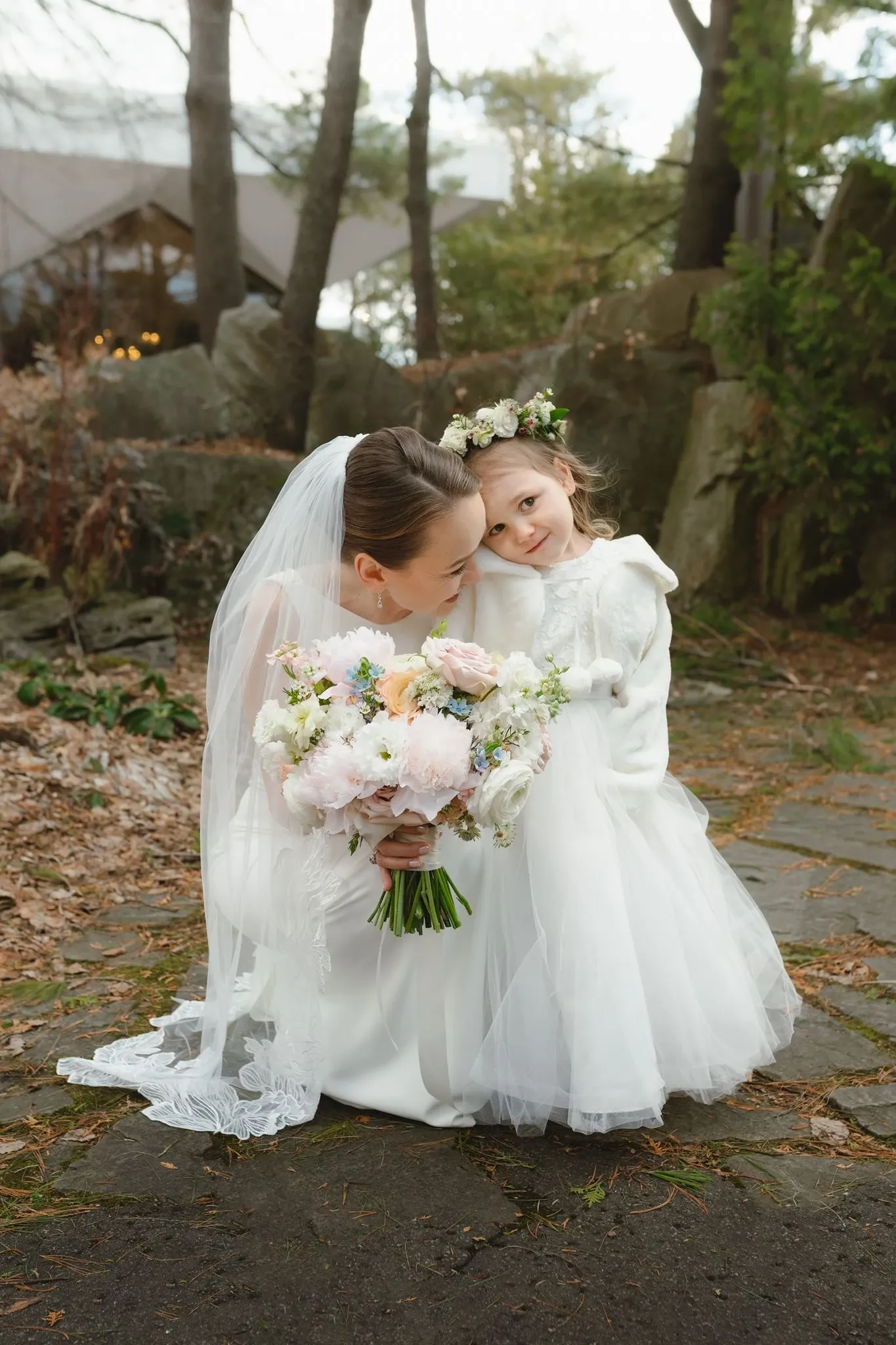 Une mariée élégamment vêtue d'une robe blanche est agenouillée sur un chemin en pierre, tenant un grand bouquet de fleurs composé de roses pâles, de pivoines blanches et de verdure. À ses côtés, une jeune fille porte une robe blanche et un manteau chaud, avec une couronne de fleurs sur la tête. Les deux sourient tendrement, entourées par un cadre naturel agrémenté de rochers et d'arbres. Cette image, capturée lors d'un mariage en plein air, met en avant l'émotion et la beauté de l'instant, idéalement illustrée pour la catégorie des fleuristes de mariage sur le site Mariage Elegant au Canada.