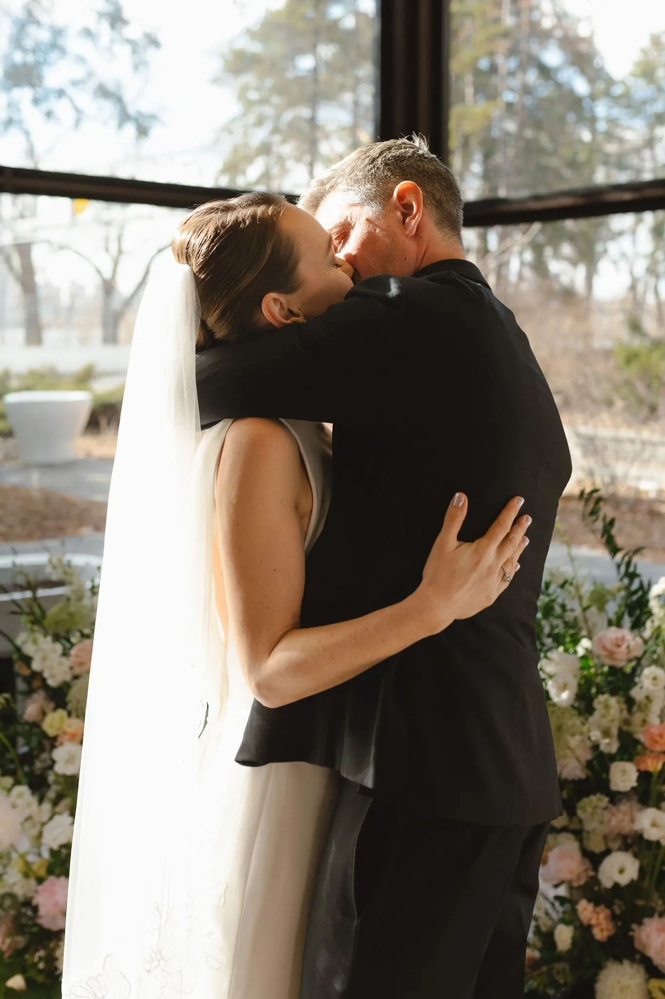 A newlywed couple shares a romantic kiss in front of a beautifully arranged floral display. The bride is wearing an elegant white wedding gown with a long veil, while the groom is in a classic black suit. The indoor setting is filled with natural light, highlighting the lush greenery and soft pastel flowers, which add a touch of elegance and romance to the scene. The floral arrangement in the background, crafted by a wedding florist, enhances the intimate and celebratory atmosphere of the moment.