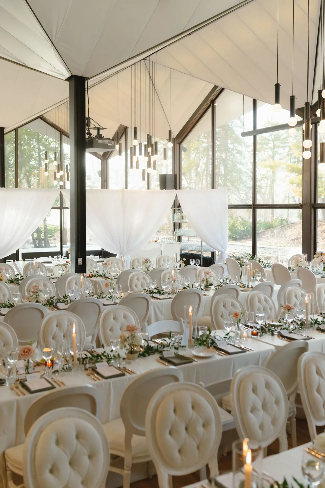 The image depicts an elegantly decorated wedding reception venue featuring a sophisticated floral arrangement. The space is set with round tables adorned with white linens and delicately padded chairs. Each table is beautifully decorated with long, lush garlands of greenery interspersed with pink and white flowers. Elegant taper candles in glass holders add a soft, romantic glow to the setting. Large windows provide ample natural light, and modern pendant lights hang from the ceiling. Sheer white drapes create a gentle, intimate ambiance, enhancing the luxurious feel of the venue. The setting is perfect for a stylish wedding celebration, exemplifying the work of a talented wedding florist in Canada, as featured in Mariage Elegant's wedding planning and vendor listing.
