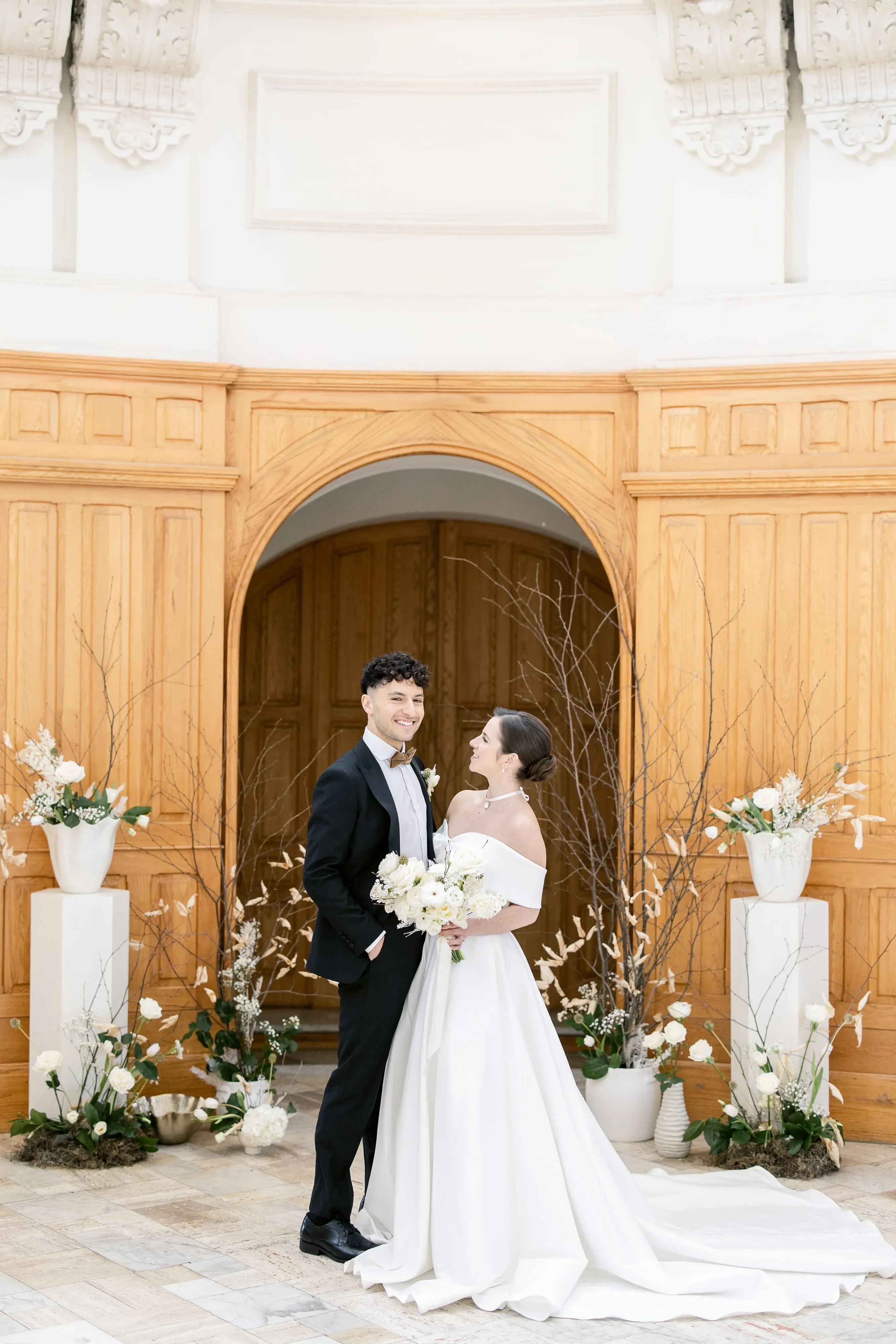 A bride and groom stand in an elegant wedding venue, gazing at each other with smiles. The bride is dressed in an off-the-shoulder white gown and is holding a bouquet of white flowers, while the groom wears a classic black tuxedo with a white shirt. They are framed by a beautifully arranged floral display featuring tall white vases with white flowers and delicate twigs, enhancing the romantic atmosphere. The background showcases ornate wooden paneling and an arched doorway, contributing to the sophisticated setting ideal for a wedding ceremony or photography session.