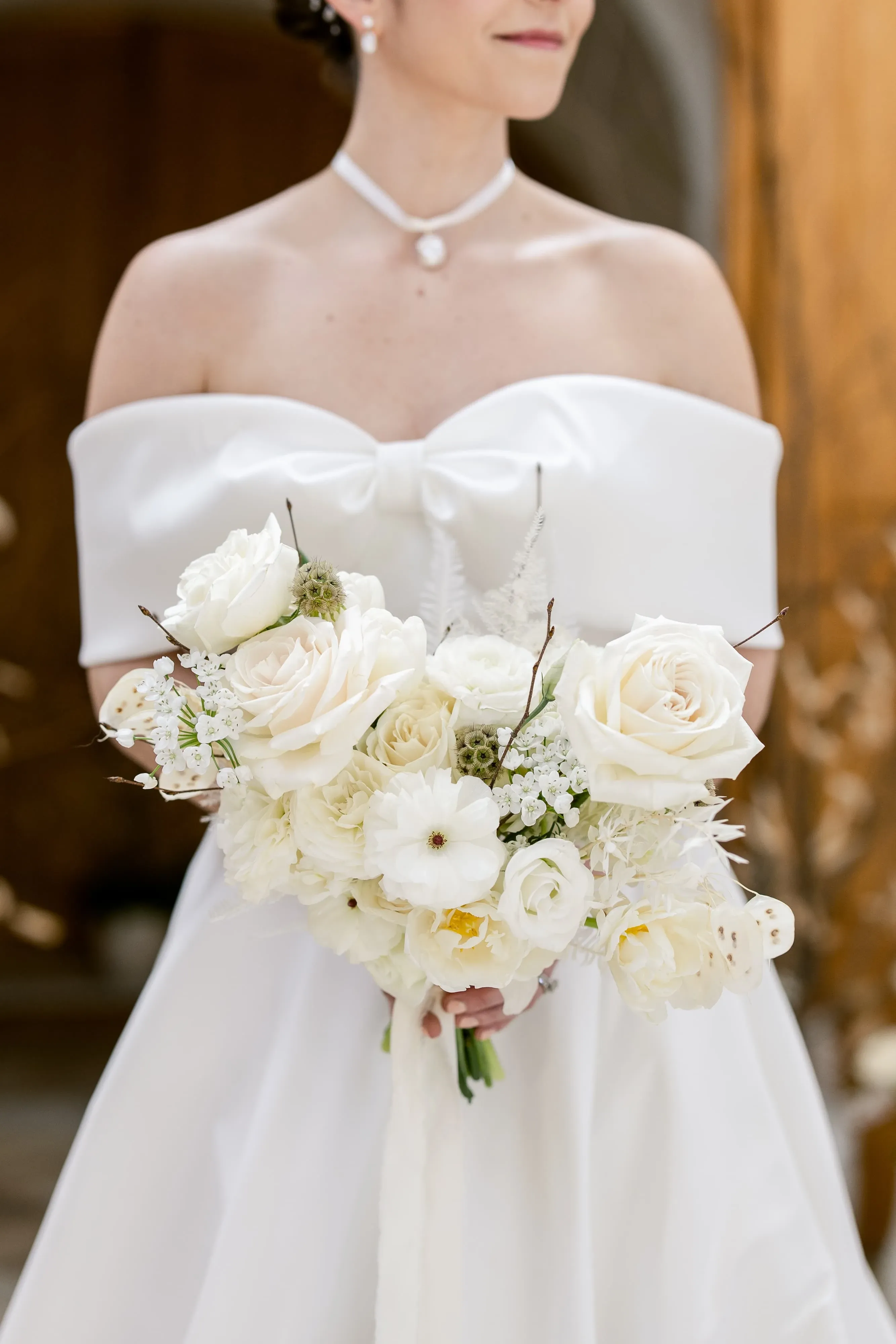 A bride is elegantly dressed in an off-the-shoulder, satin wedding gown, showcasing a classic yet modern style. She holds a sophisticated bouquet predominantly featuring white roses, delicately accented with smaller white blooms and lush greenery. The arrangement exudes a timeless beauty, perfect for an elegant wedding setting. The background is softly blurred, emphasizing the focus on the bride and her exquisite floral arrangement. The image captures a moment of refined elegance, suitable for a wedding florist listing on Mariage Elegant.