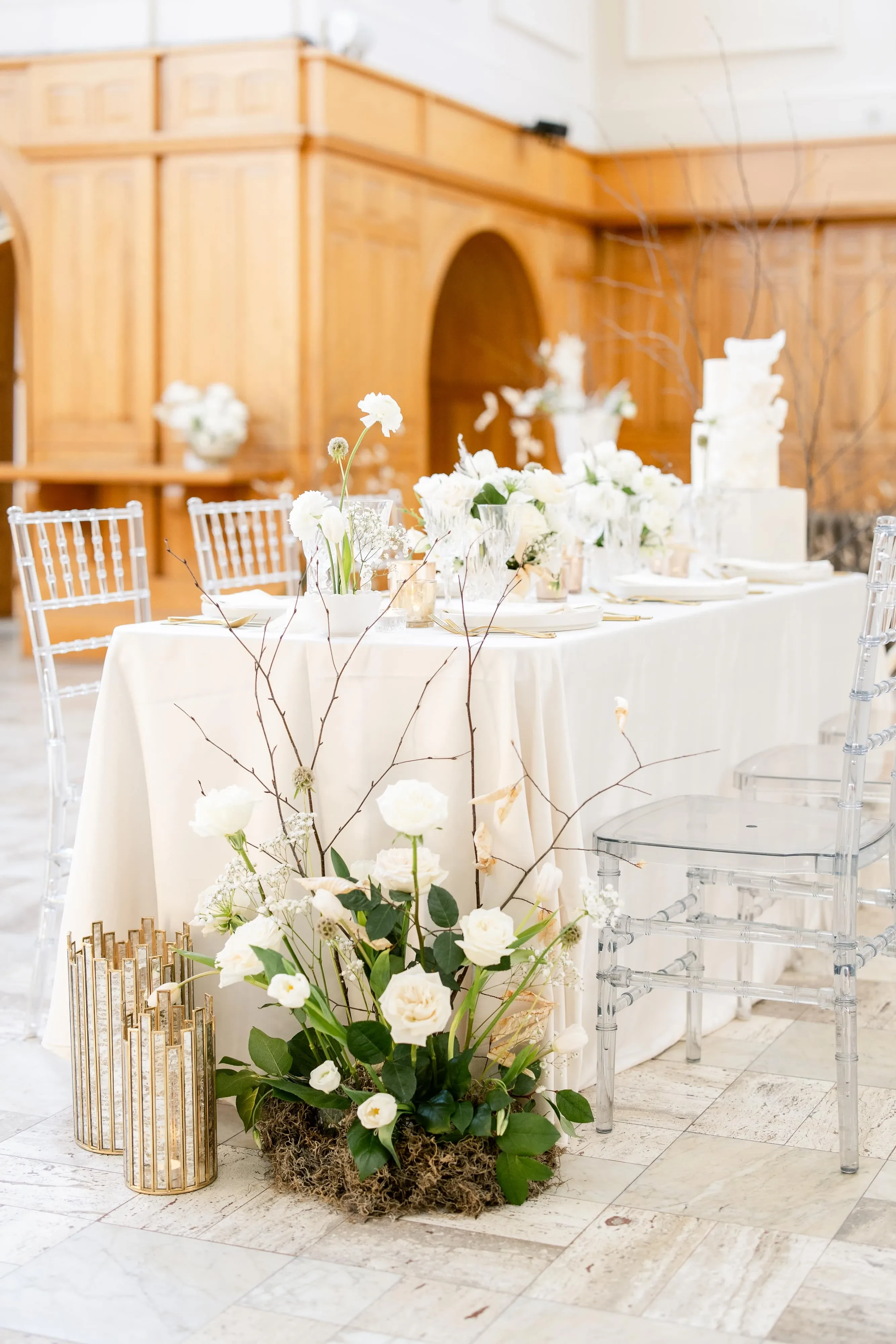 The image showcases a beautifully arranged wedding reception table set up in a sophisticated indoor venue, highlighting the work of a wedding florist. The table is adorned with an elegant white tablecloth and features a centerpiece composed of white roses and delicate greenery, creating a fresh and elegant look. Transparent chairs are placed around the table, enhancing the modern and classy ambiance. In the foreground, a refined floral arrangement sits on the floor, featuring white roses, greenery, and decorative gold elements that add a touch of luxury. The background reveals wooden architectural details, adding warmth and contrast to the bright and airy setting. The overall composition reflects a timeless and elegant wedding theme, ideal for couples seeking a stylish and sophisticated event in Canada.