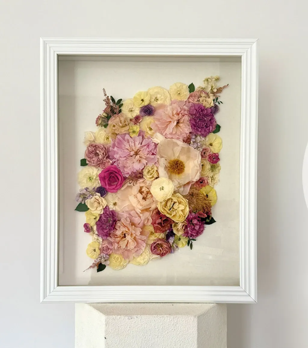 A beautifully arranged collection of dried and pressed flowers is artfully displayed within a white, framed shadow box. The assortment includes delicate roses and other blooms in soft shades of pink, yellow, and cream, suggesting a romantic and elegant theme suitable for weddings. The arrangement is tastefully mounted against a light background, highlighting each flower's intricate details and textures. This floral art piece would be an ideal decorative element for a wedding, offering a timeless and preserved botanical aesthetic that adds charm and sophistication to any venue.