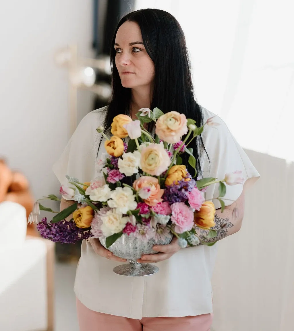 A professional florist with long dark hair is holding an elegant floral arrangement in a clear pedestal vase. The bouquet is a vibrant mix of yellow roses, pale peach ranunculus, pink carnations, and lavender sprigs, accented with greenery. The setting appears bright and airy, suitable for a wedding or event consultation. The florist is dressed in a casual white blouse and pink pants, standing beside a softly-lit interior, which lends a welcoming and sophisticated atmosphere. This image is suitable for featuring a wedding florist vendor on a wedding planning website like Mariage Elegant in Canada.