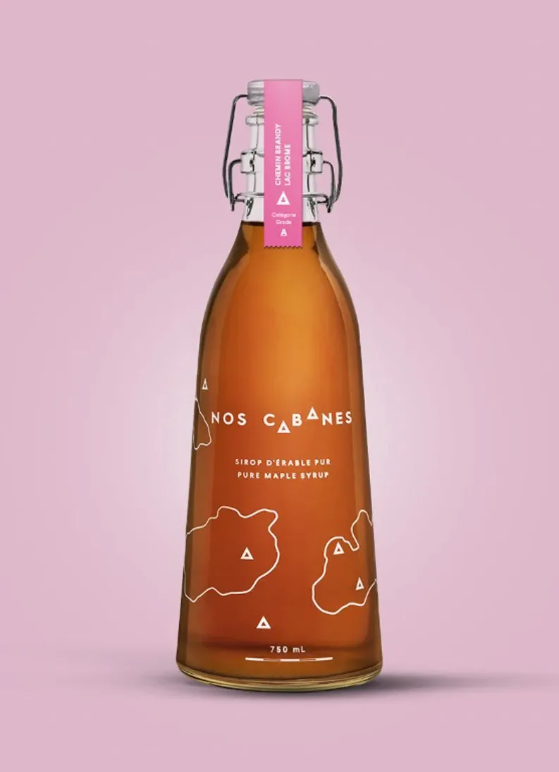 Une bouteille en verre de 750 mL de sirop d'érable pur 'Nos Cabanes' est posée contre un fond rose. La bouteille est dotée d'un bouchon mécanique et d'une étiquette rose indiquant 'Cadeau Classique Mariage Elegant'. Le sirop d'érable, produit artisanal, est présenté comme une option de cadeau de mariage raffinée et typique du Canada, idéale pour figurer dans une liste de cadeaux de mariage haut de gamme.