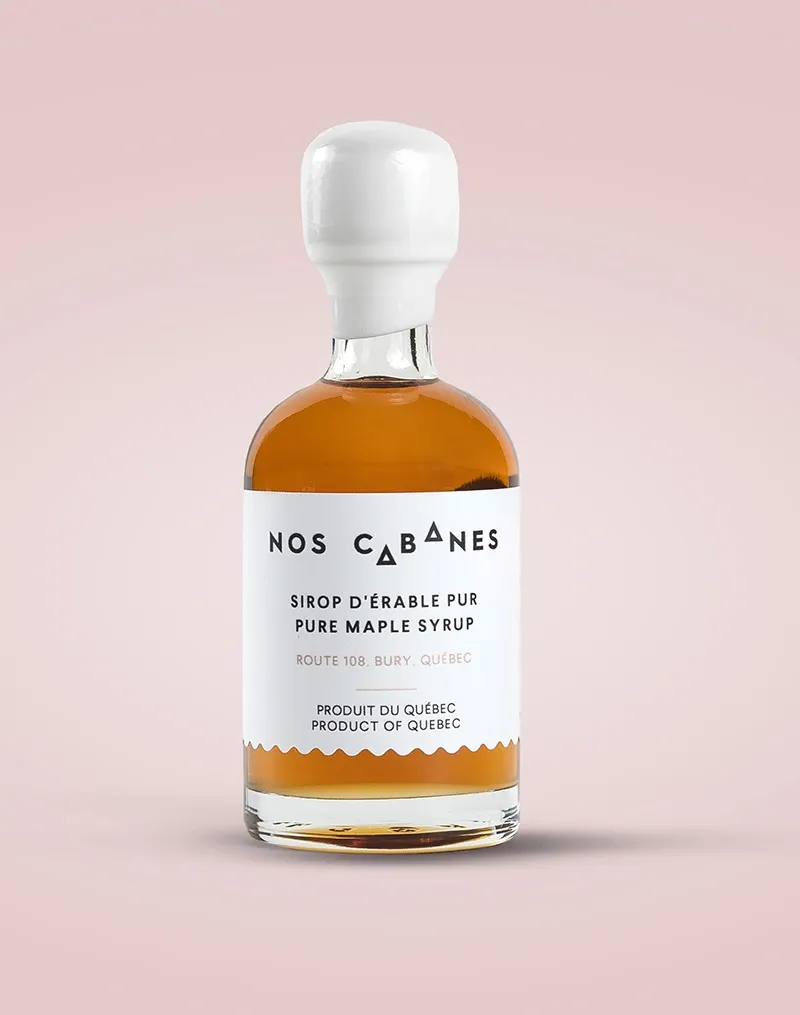 The image features a glass bottle of pure maple syrup from the brand 'Nos Cabanes,' set against a soft pink background. The bottle has a clean, modern design with a white cap. The label, predominantly white, displays text in both English and French, highlighting the product's origin from Quebec, Canada. This maple syrup bottle is presented as a potential wedding favor option, complementing elegant wedding themes and emphasizing local Canadian products.