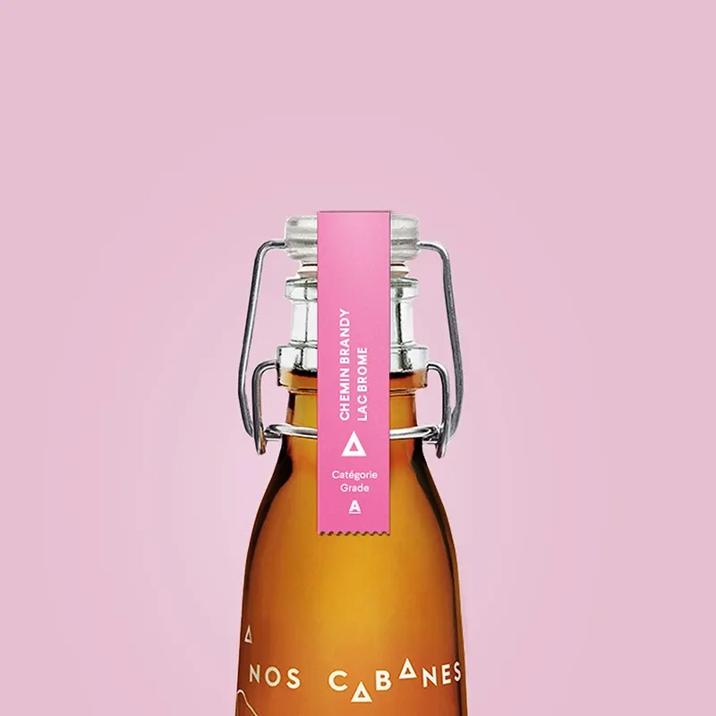 A glass bottle filled with amber liquid is prominently displayed against a soft pink background. The bottle has a secure, swing-top closure sealed with a metal clasp. Attached to the top is a pink label with white text that reads 'CHEMIN BRANDY LA CABANE' and indicates a 'Catégorie Grade A.' The bottle is a high-quality option for wedding favors, offering a charming and sophisticated gift for guests. This elegant presentation is ideal for adding a touch of luxury to any wedding celebration, enhancing the event's overall aesthetic.