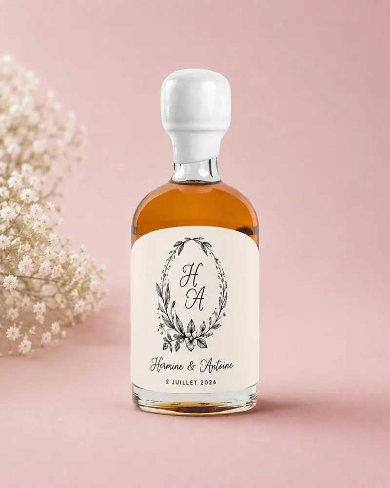 A small glass bottle containing amber-colored liquid, likely a personalized wedding favor, is set against a soft pink background. The bottle features a custom label with an elegant monogram of the initials 'H A' surrounded by a delicate floral wreath design. Below the monogram, the names 'Hermine & Antoine' and the date '2 Juillet 2026' are printed, suggesting a wedding or event date. To the left of the bottle is a sprig of baby's breath flowers, enhancing the romantic and sophisticated aesthetic suitable for wedding celebrations.