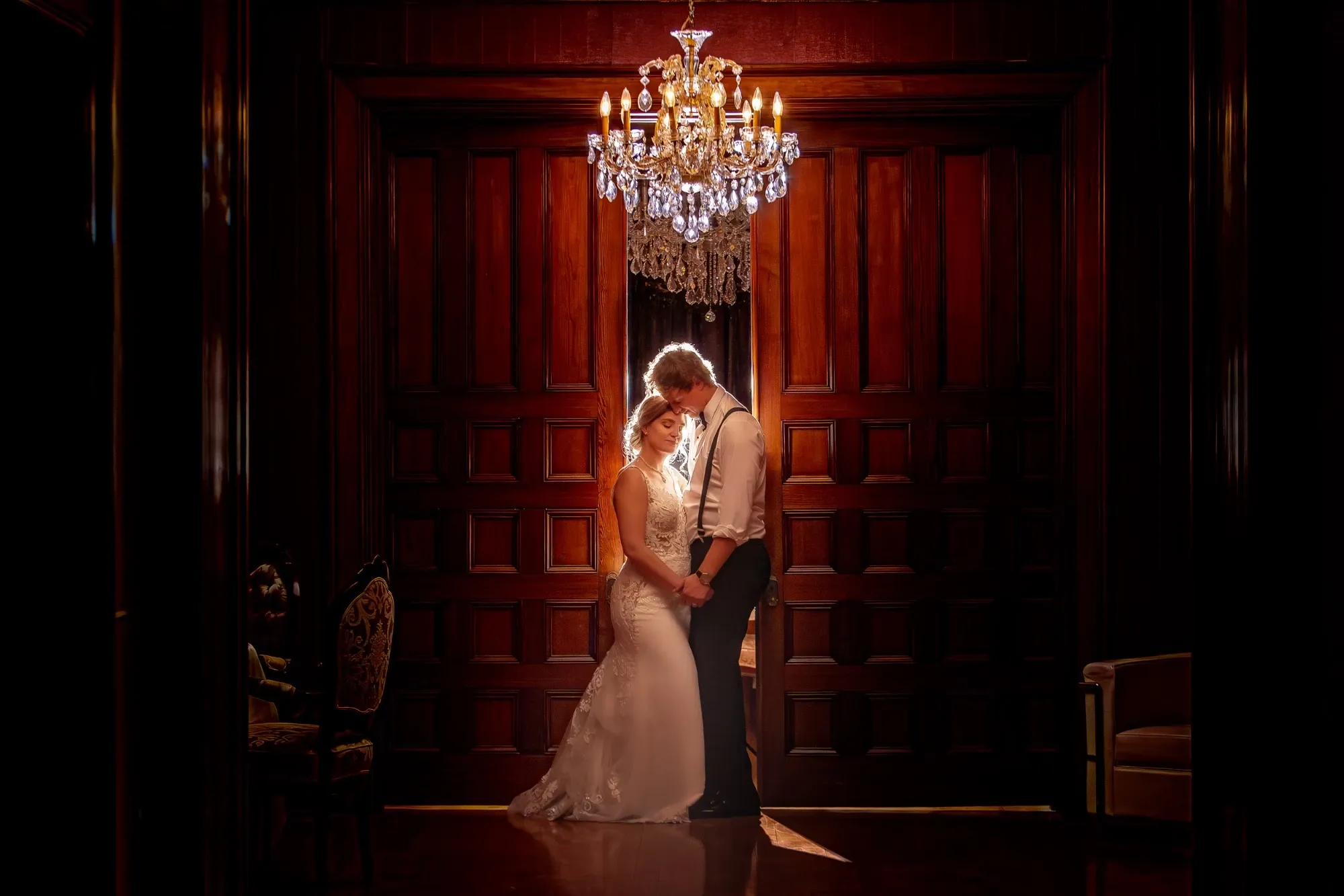 The image showcases a romantic wedding scene captured by Inhouse Studios Photography, a vendor specializing in wedding photography. In a dimly lit, elegant room with rich wooden paneling, a bride and groom share an intimate moment. They stand beneath a sparkling chandelier, creating a warm and inviting atmosphere. The bride is wearing a classic white gown with intricate lace details, and the groom is dressed in formal attire with suspenders. Their gentle embrace and the soft lighting highlight the emotion and elegance of the occasion.