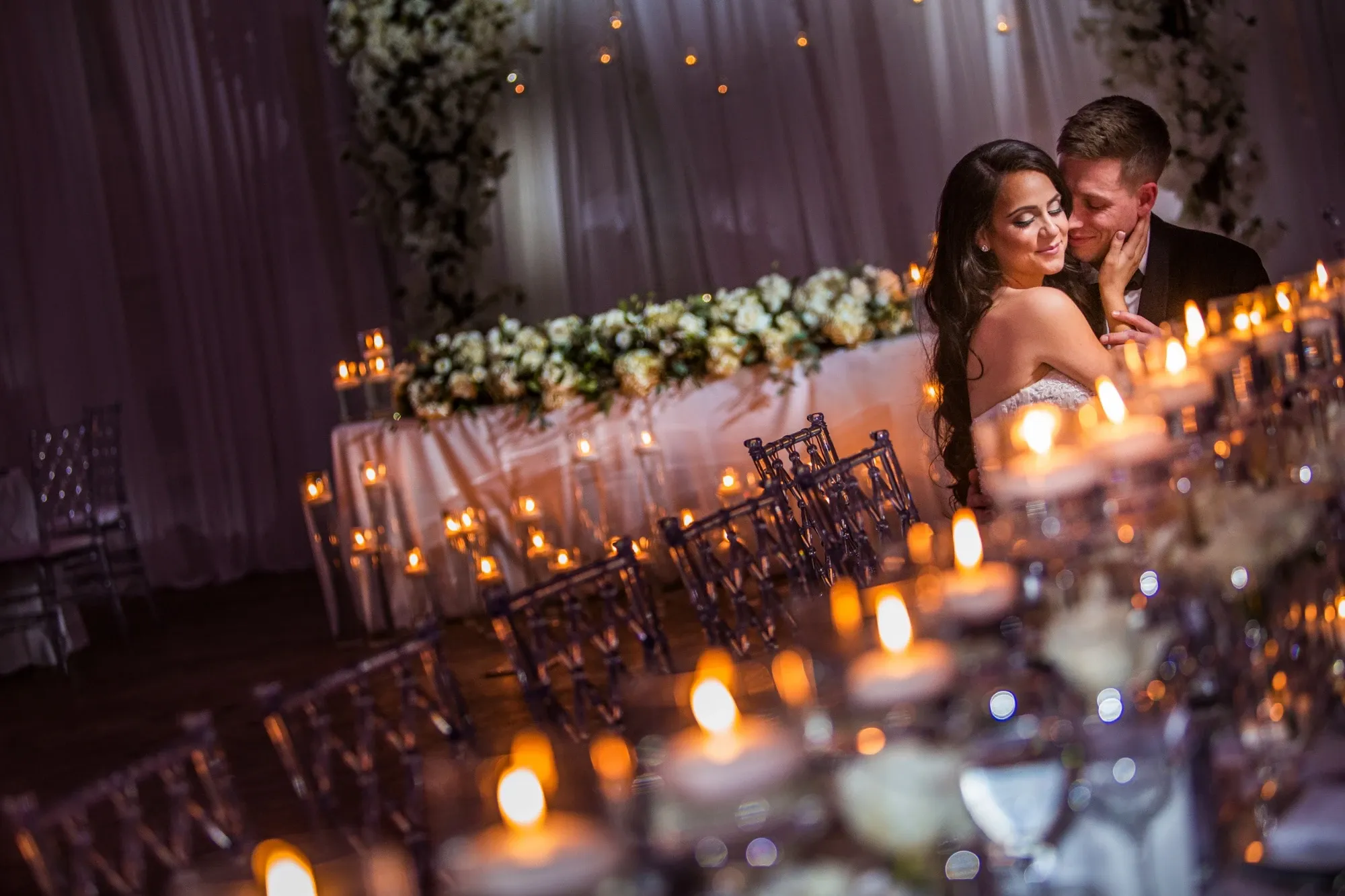 A romantic evening wedding reception photograph captured by Inhouse Studios Photography, exemplifying their expertise in wedding photography. The image features a newlywed couple lovingly embracing amidst a beautifully decorated setting. The atmospheric glow of numerous candlelit tables, elegantly arranged with florals, adds a warm and intimate ambiance. In the background, a stunning floral arch frames the couple, highlighting the festive environment suited for a wedding celebration.