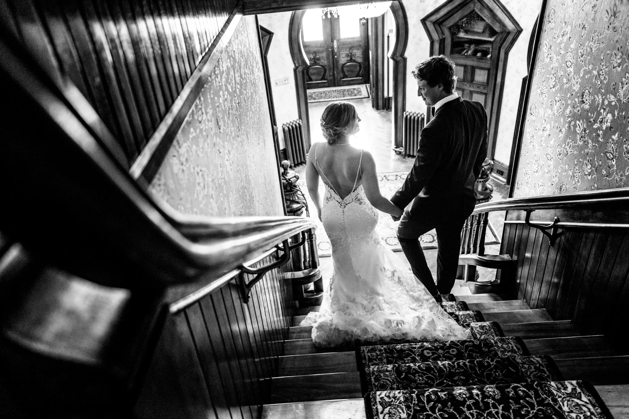 A bride and groom descend an ornate wooden staircase in an elegant, historic venue. The bride is wearing a stunning lace wedding dress with a long train, while the groom is in a classic dark suit. They are captured from behind, highlighting the intricate details of the bride's gown. Soft light filters through a window, creating a warm, romantic ambiance. This image showcases the sophisticated and artistic style of Inhouse Studios Photography, a vendor specializing in wedding photography, exemplifying their ability to capture intimate, beautiful moments for weddings in Canada.