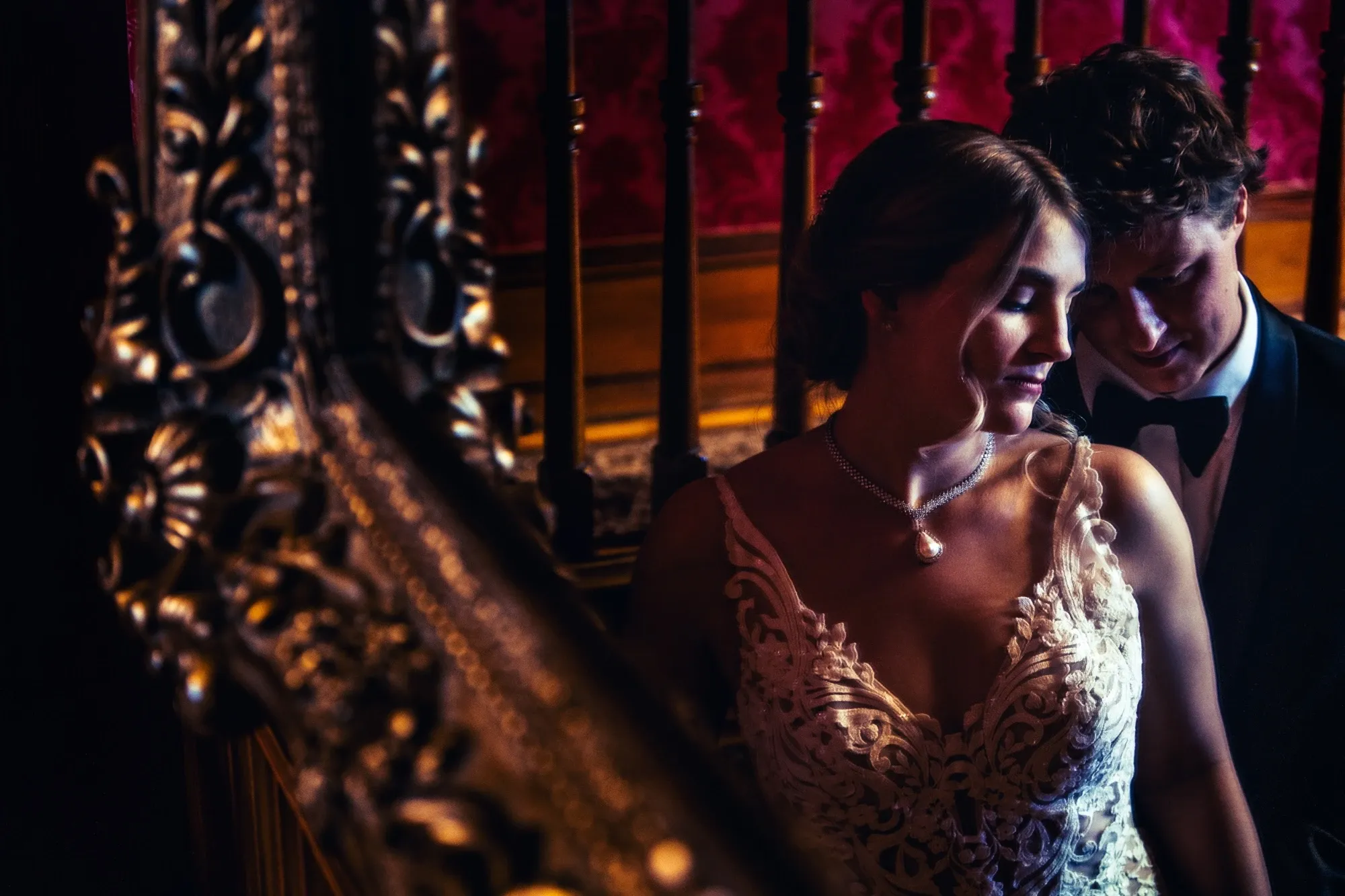 Inhouse Studios Photography captures an intimate moment of a bride and groom in a dimly lit staircase setting. The bride is wearing an intricate lace wedding gown with a delicate pearl necklace that complements her elegant look. The groom is dressed in a classic black tuxedo, gently leaning towards the bride. The setting features a luxurious wooden banister and rich, red wallpaper that add a romantic and timeless atmosphere to the scene. This image highlights the wedding photography expertise of Inhouse Studios Photography, showcasing a blend of style and emotion perfect for couples planning their special day with Mariage Elegant in Canada.