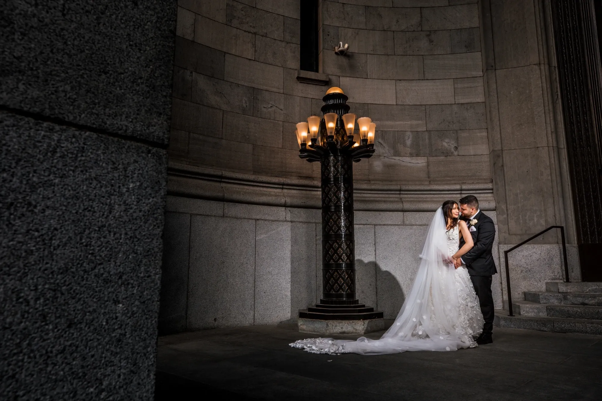 In this elegant wedding photography image by Inhouse Studios Photography, a bride and groom are seen embracing under a grand, ornate lamp post in a sophisticated stone setting, highlighting the romantic and intimate mood of their special day. The bride is wearing a stunning, white lace gown with a flowing veil cascading to the floor, while the groom is in a classic dark suit, perfectly complementing the luxurious and timeless backdrop. This photo showcases the artistry of wedding photography, capturing a tender moment against the dramatic architecture, ideal for those seeking timeless elegance in their wedding memories.