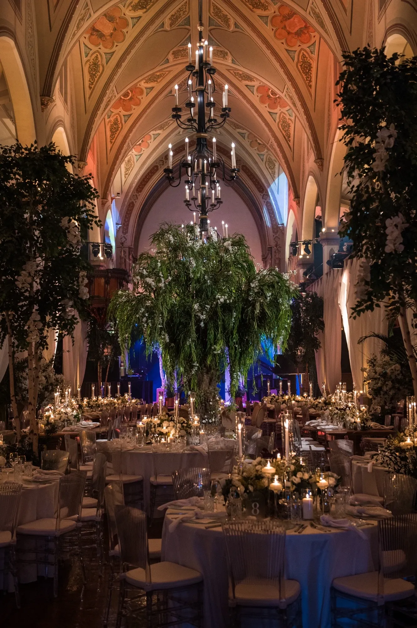 The image showcases a beautifully decorated wedding reception venue captured by Inhouse Studios Photography, specializing in wedding photography. The elegant setting features grand arched ceilings adorned with intricate designs. A striking central piece is a towering arrangement of lush greenery, complemented by ambient candlelight on the tables below. The tables are set with fine china and crystal glassware, surrounded by clear chairs, creating a sophisticated atmosphere. The overall scene exudes a romantic and luxurious vibe, ideal for an unforgettable wedding celebration.